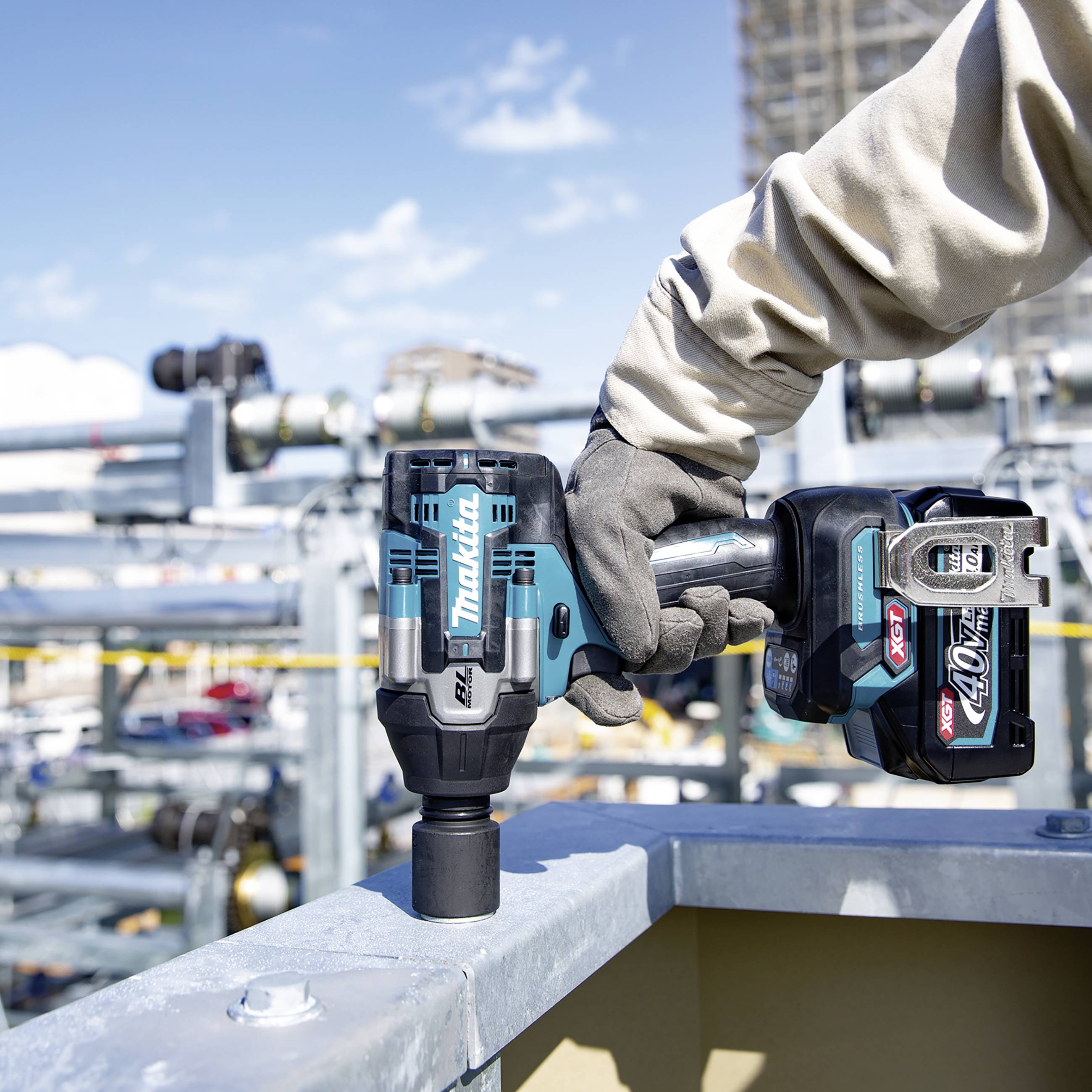Person in Arbeitskleidung benutzt eine Makita-Akku-Bohrmaschine auf einer Baustelle, umgeben von Metallstrukturen und Werkzeugen.