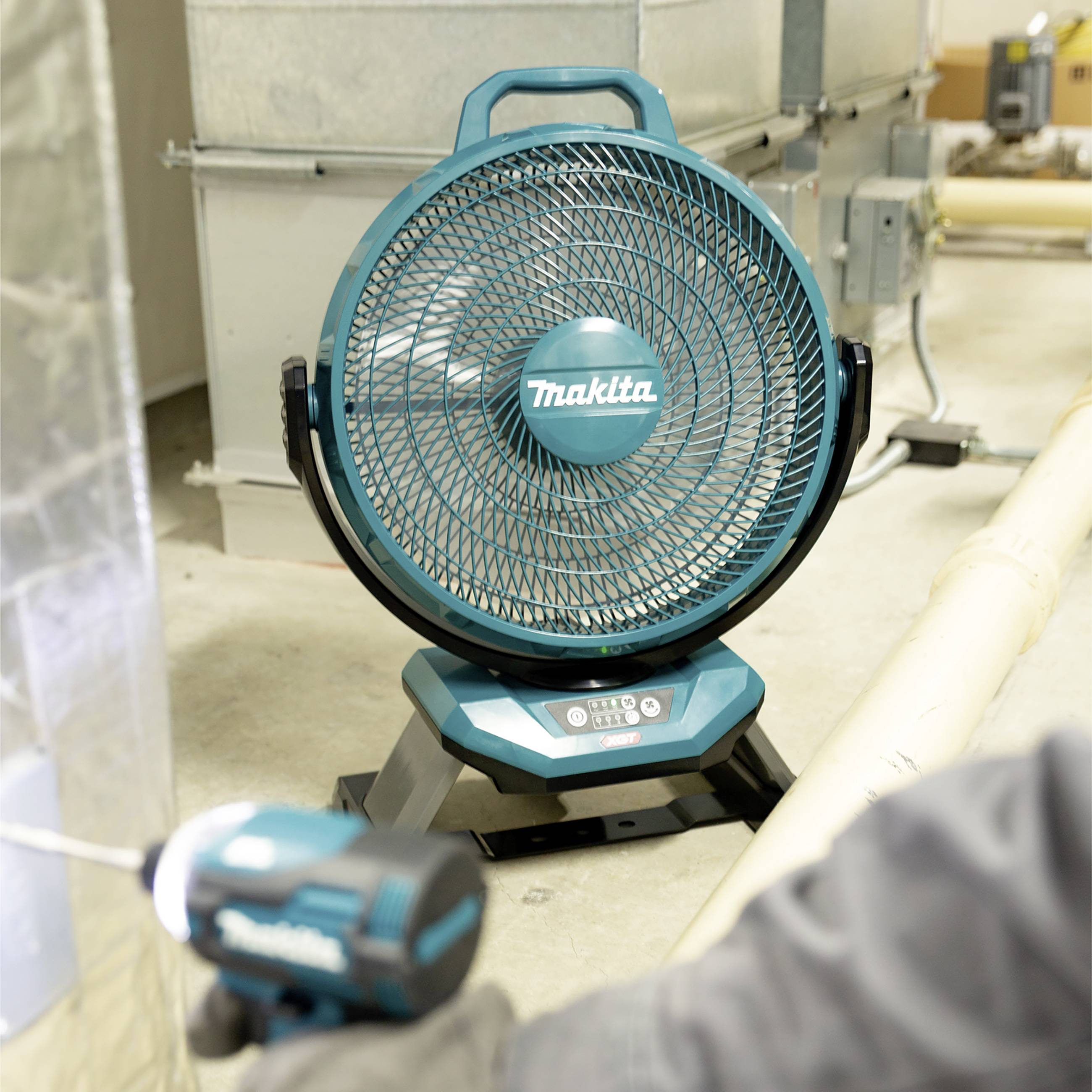 Ein blauer, tragbarer Ventilator steht in einem industriellen Raum. Im Vordergrund hält eine Person ein Werkzeug.