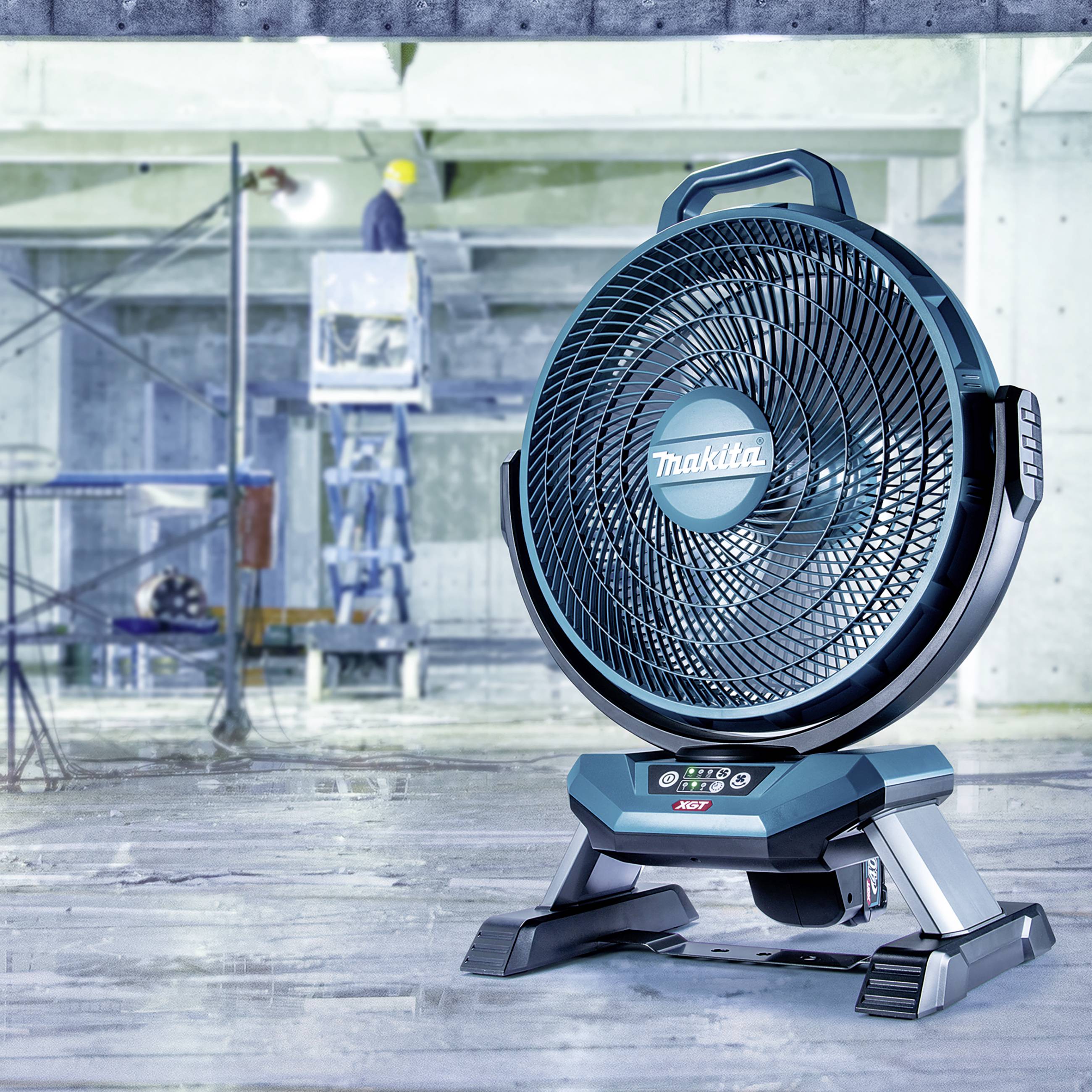 Ein blauer Ventilator steht in einem unfertigen Gebäude. Im Hintergrund arbeitet eine Person mit einem Bauhelm.