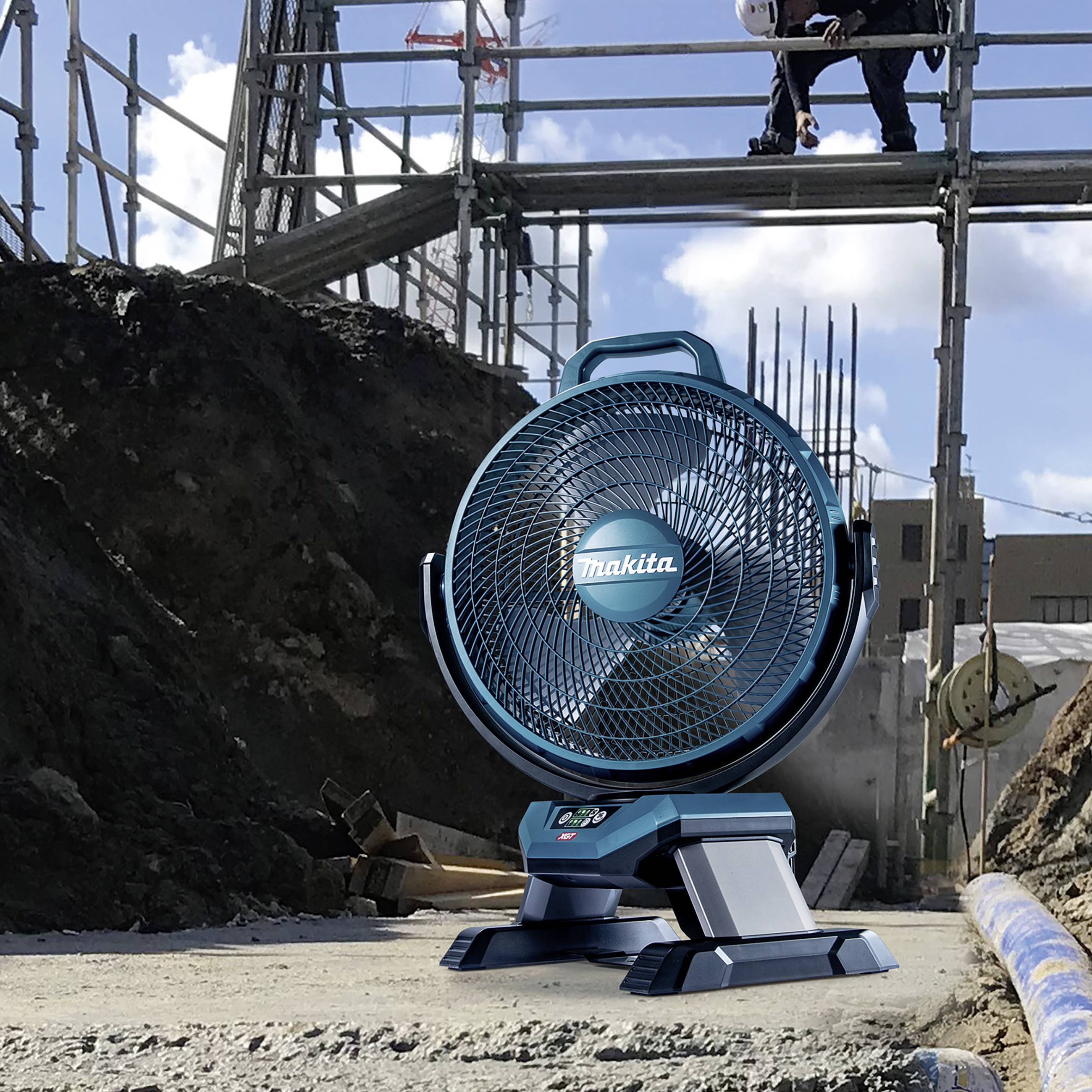 Ein Baustellenventilator steht auf dem Boden im Vordergrund. Im Hintergrund arbeiten Personen auf einem Baugerüst über einer Erdgrube.