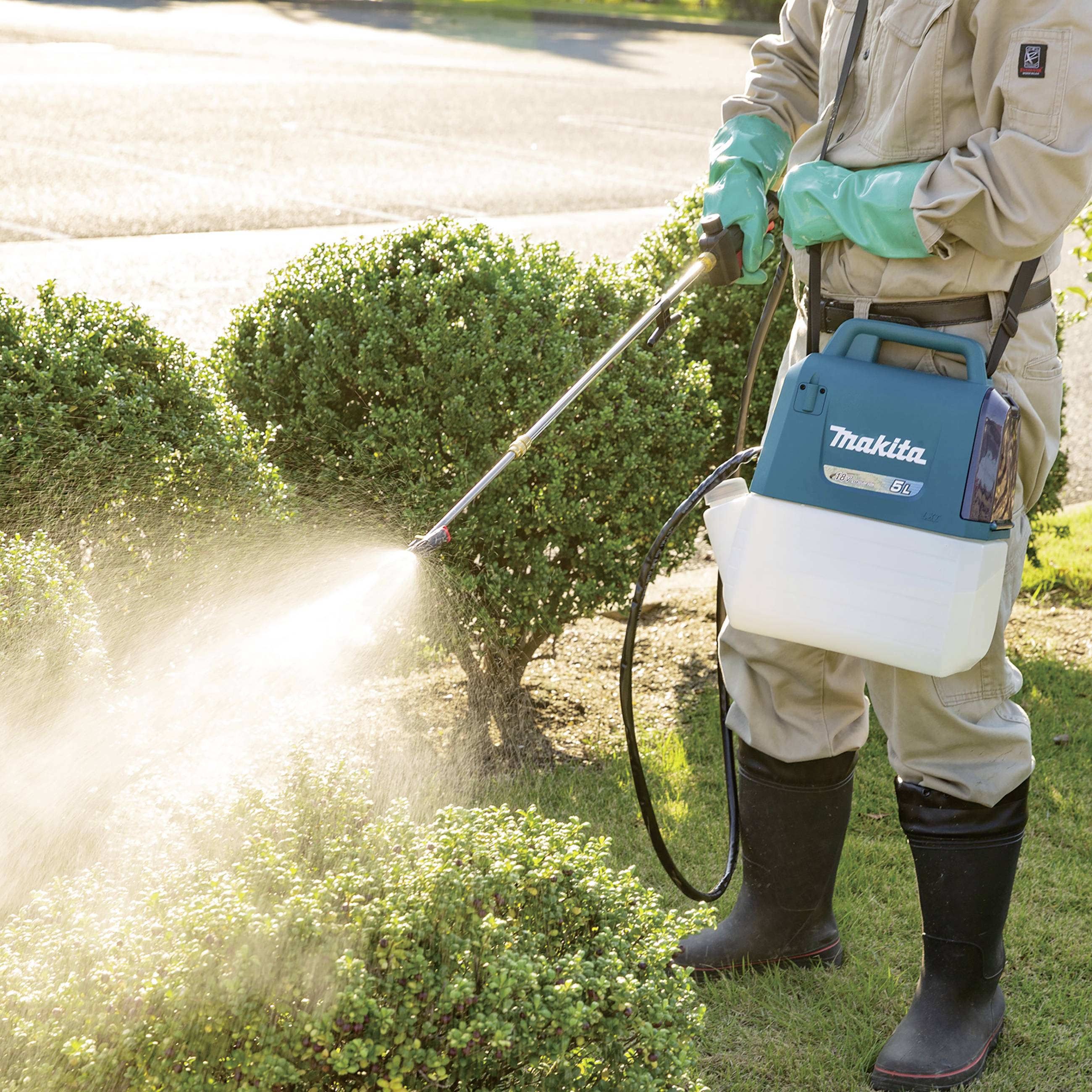 Person gießt Sträucher mit einem Sprühgerät im Garten. Sie trägt Handschuhe und Gummistiefel. Sonnenlicht scheint auf die Pflanzen.