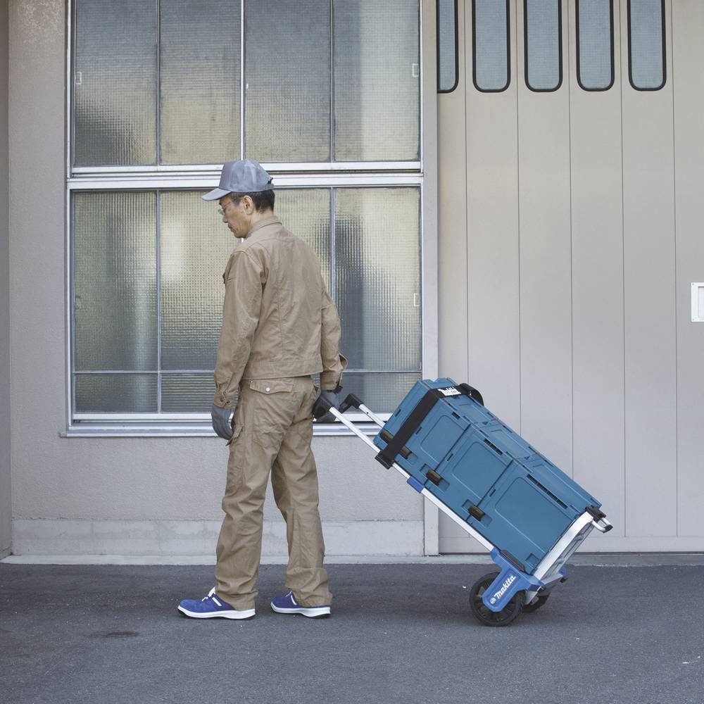 Eine Person in Arbeitskleidung zieht einen blauen Transportkarren mit Werkzeugkästen vor einem Gebäude.