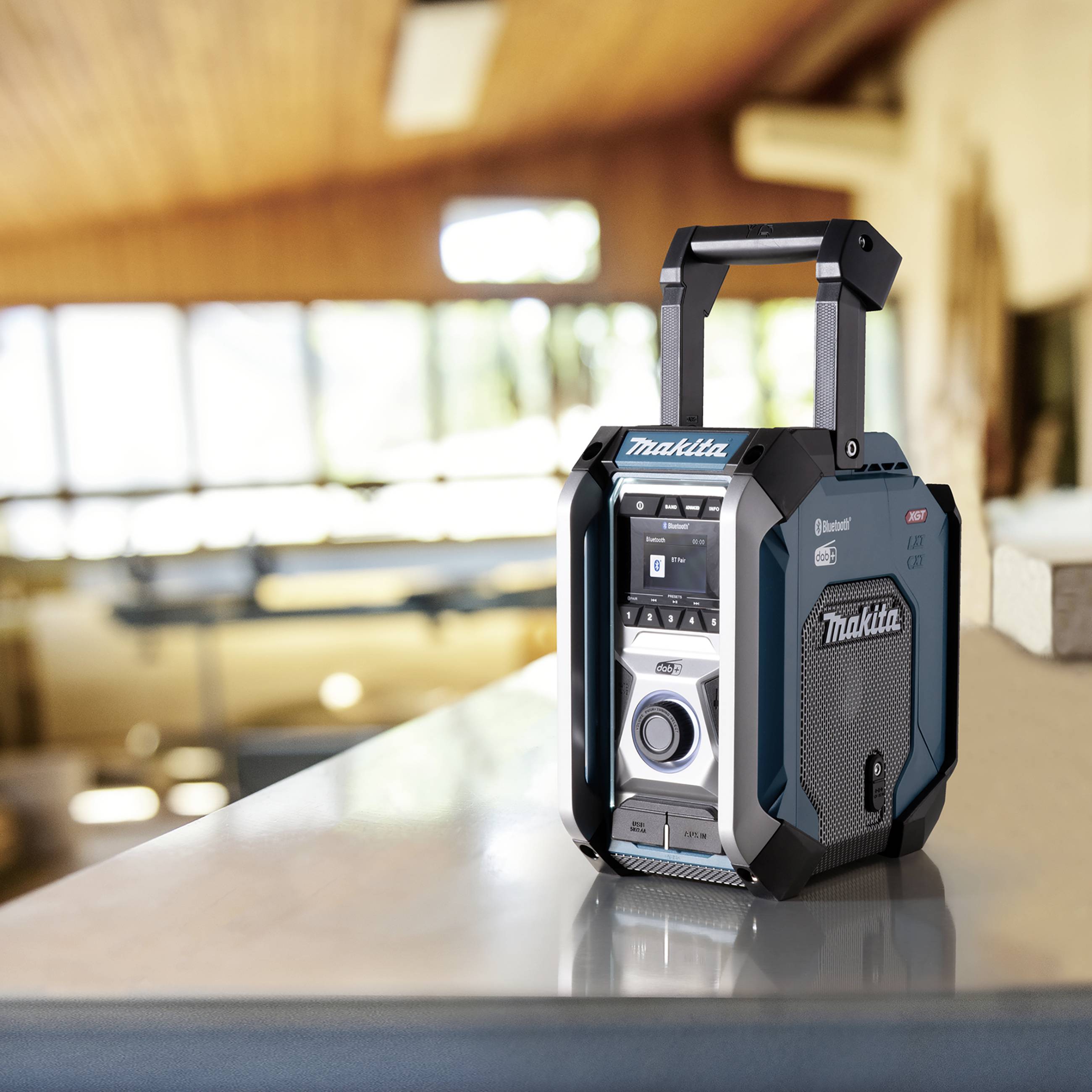 Ein tragbares Radio mit Bluetooth-Funktion der Marke Makita steht auf einem Tisch in einem hellen Raum mit Holzelementen und Fenstern.
