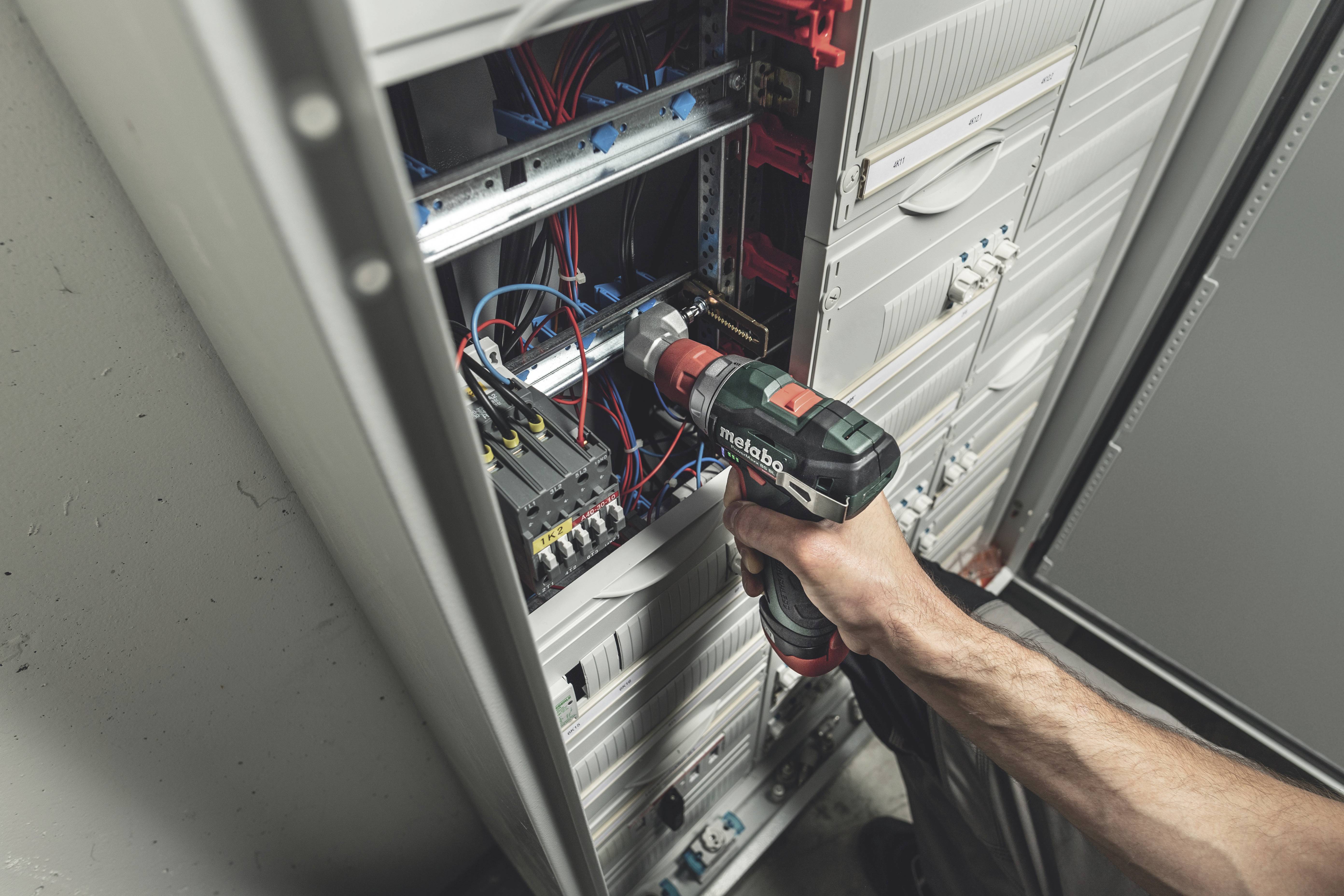 Eine Person verwendet einen Akkuschrauber, um an einem geöffneten Schaltschrank zu arbeiten, in dem viele bunte Kabel zu sehen sind.