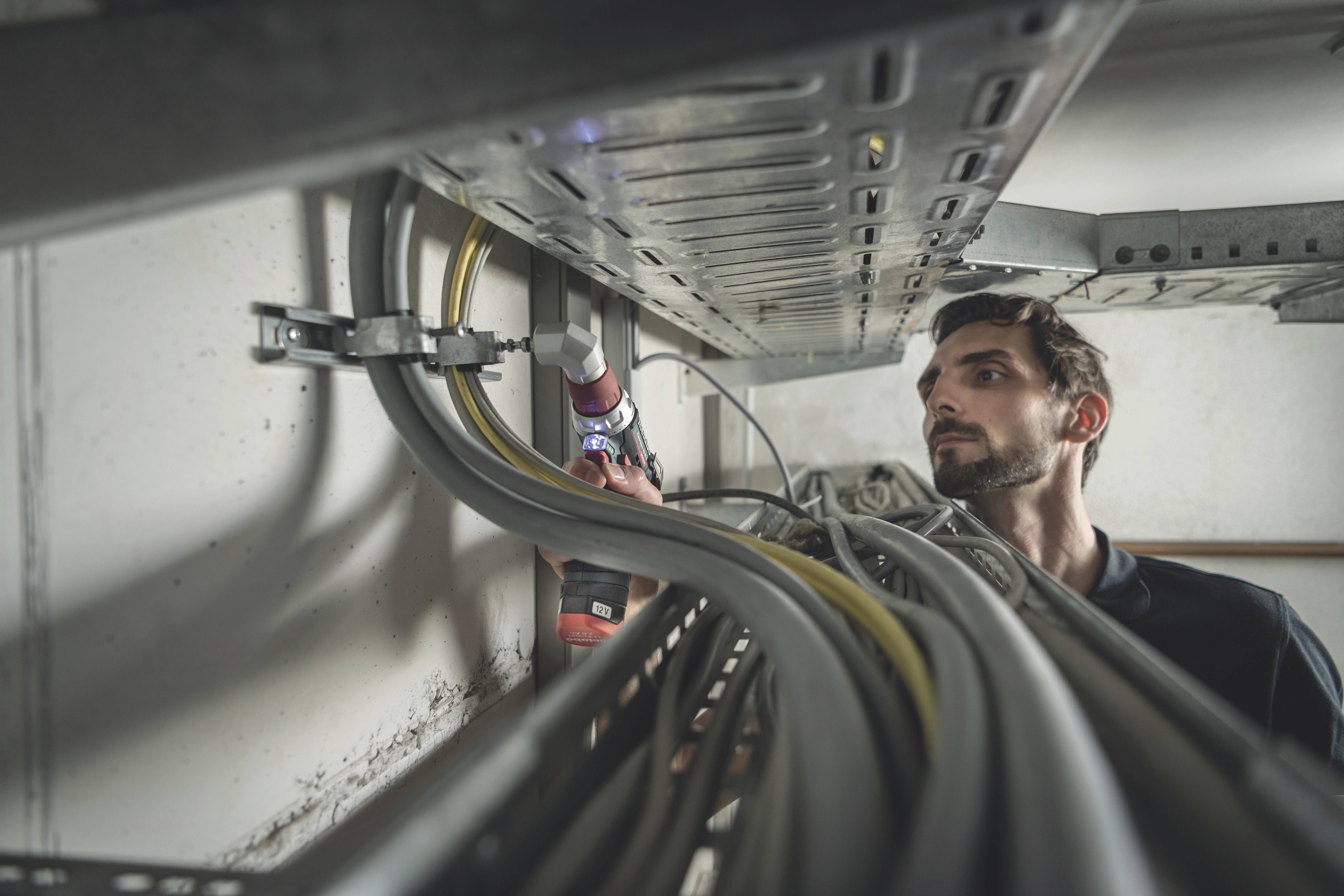 Ein Mann arbeitet an einem Kabelkanal in einem technischen Raum, dabei verwendet er ein Elektrowerkzeug. Kabel verlaufen entlang der Wand.