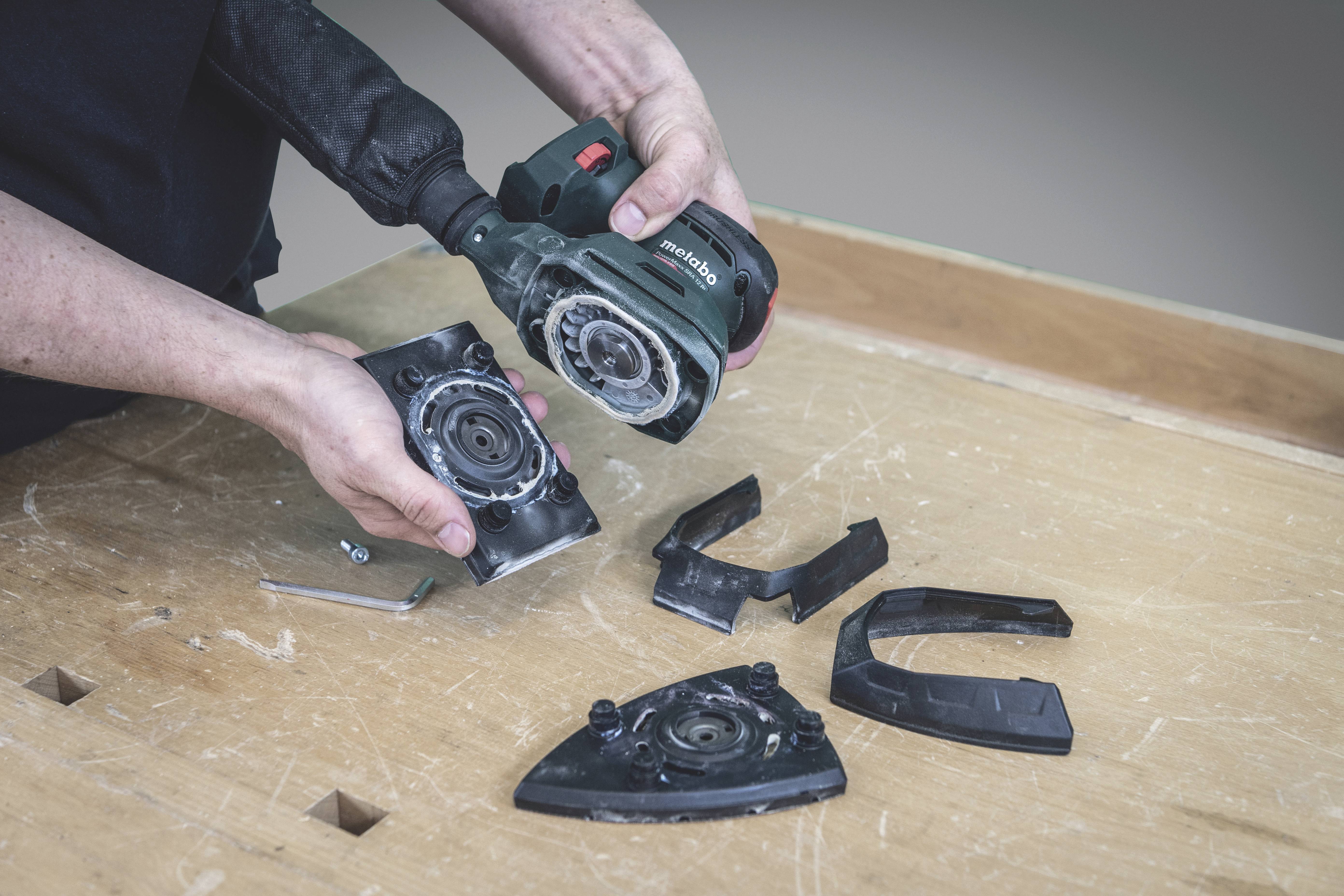 Person hält Teile einer Elektrowerkzeugmaschine auf einem Holztisch. Werkbankarbeit mit verschiedenen abnehmbaren Maschinenteilen.