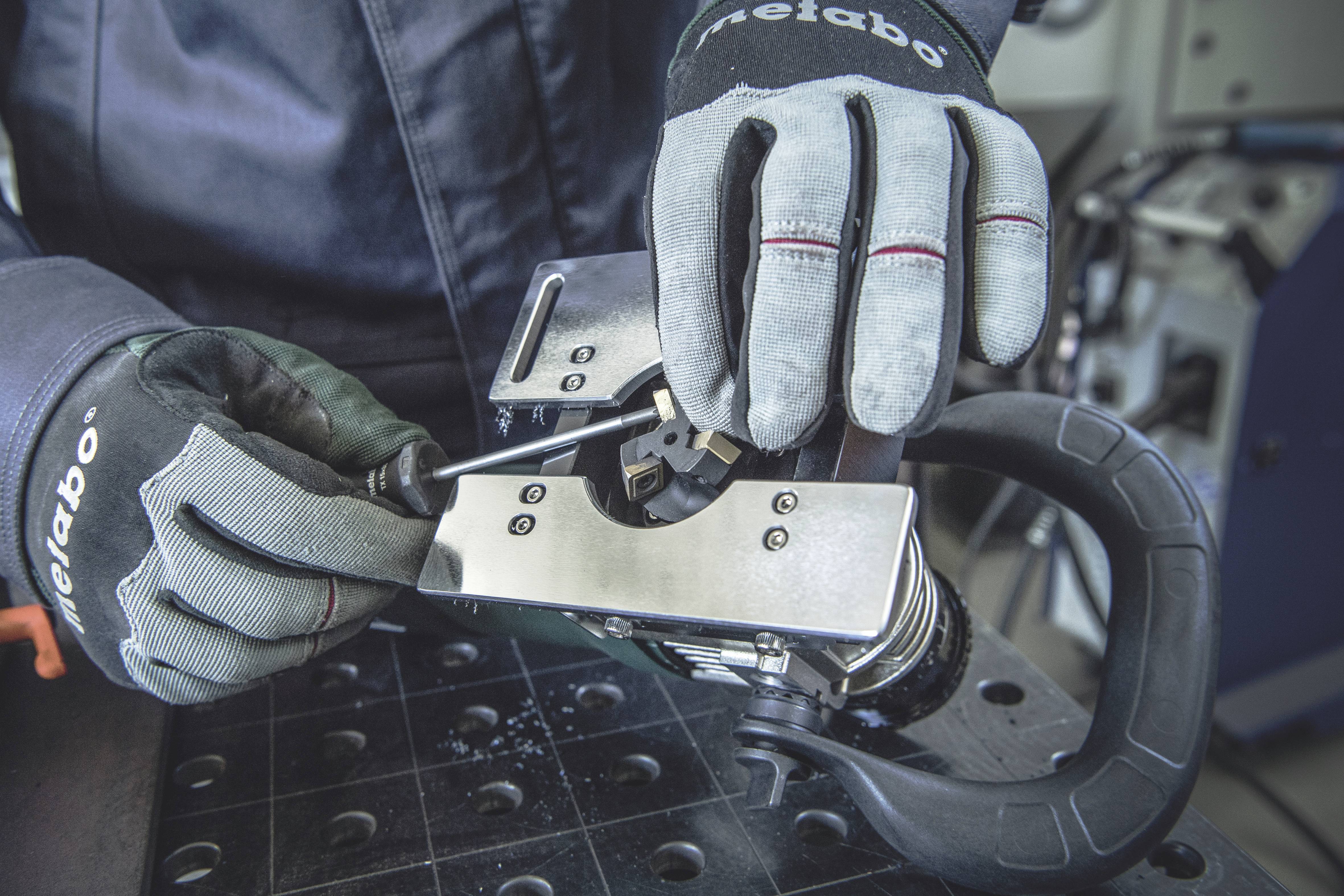 Person in Arbeitshandschuhen benutzt einen Schraubendreher zur Justierung einer Metallmaschine auf einer Werkbank.