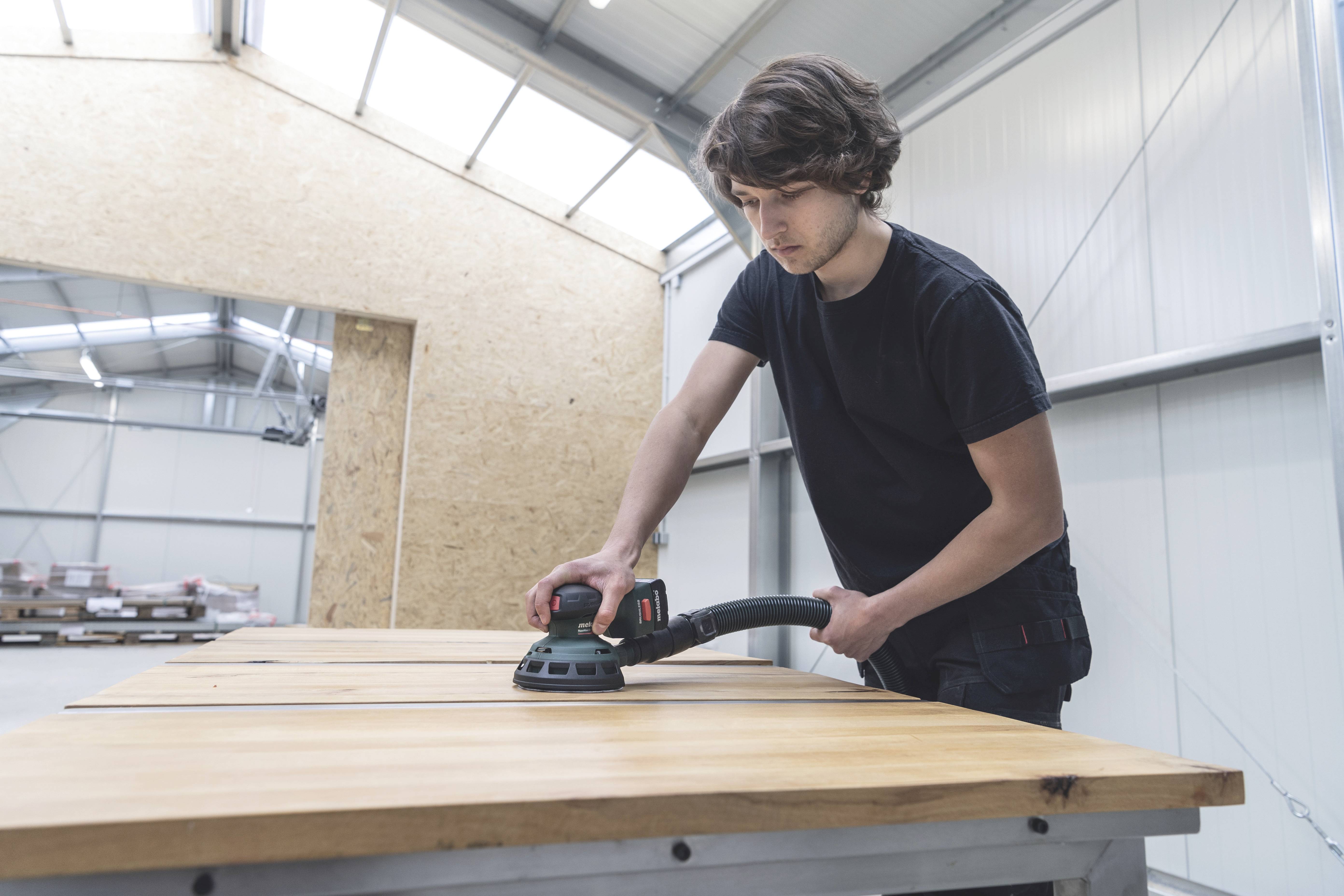 Ein Mann schleift eine große Holzplatte mit einem elektrischen Schleifgerät in einer Werkstatt.