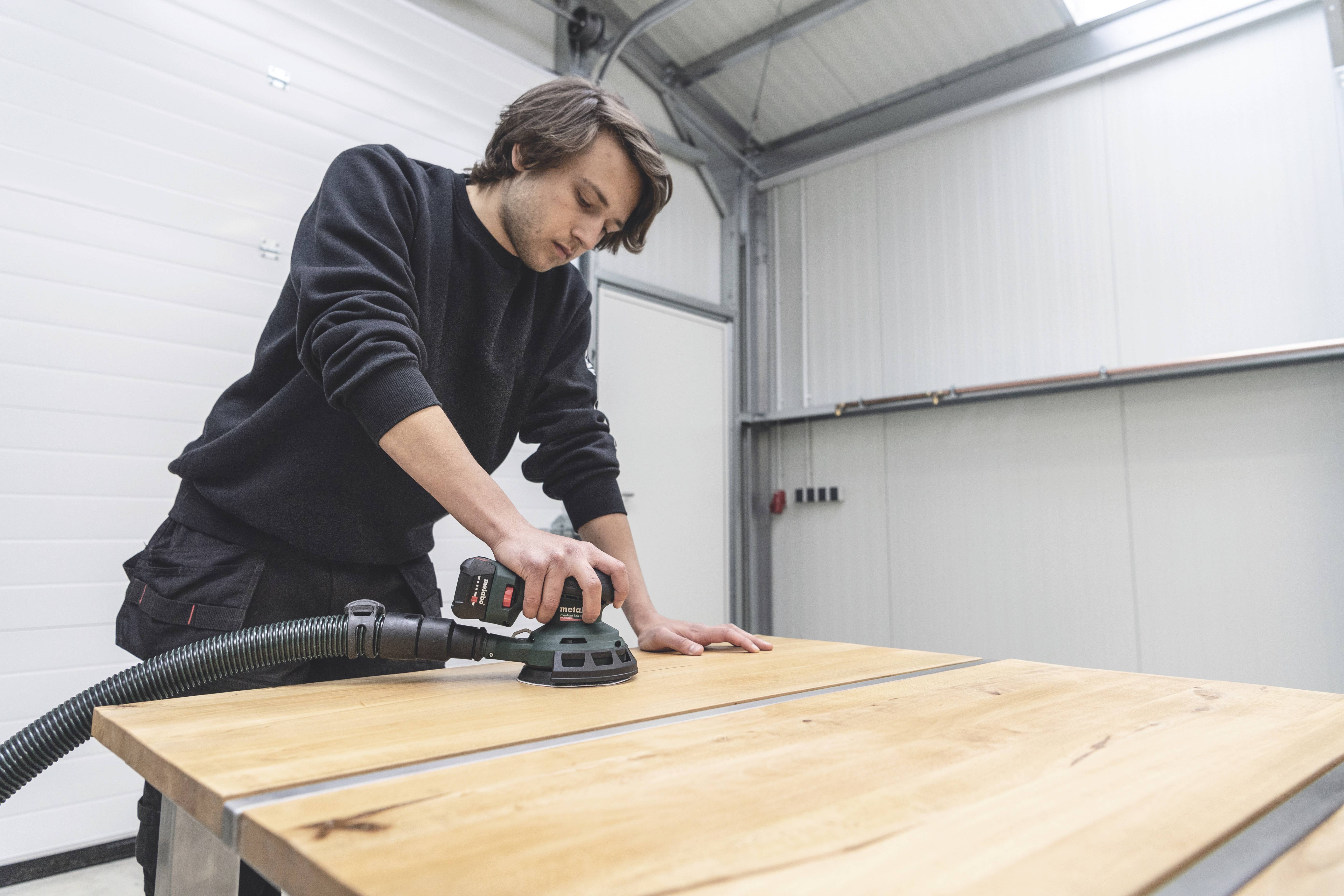 Ein Mann schleift mit einer Schleifmaschine die Oberfläche eines Holztisches in einer Werkstatt.