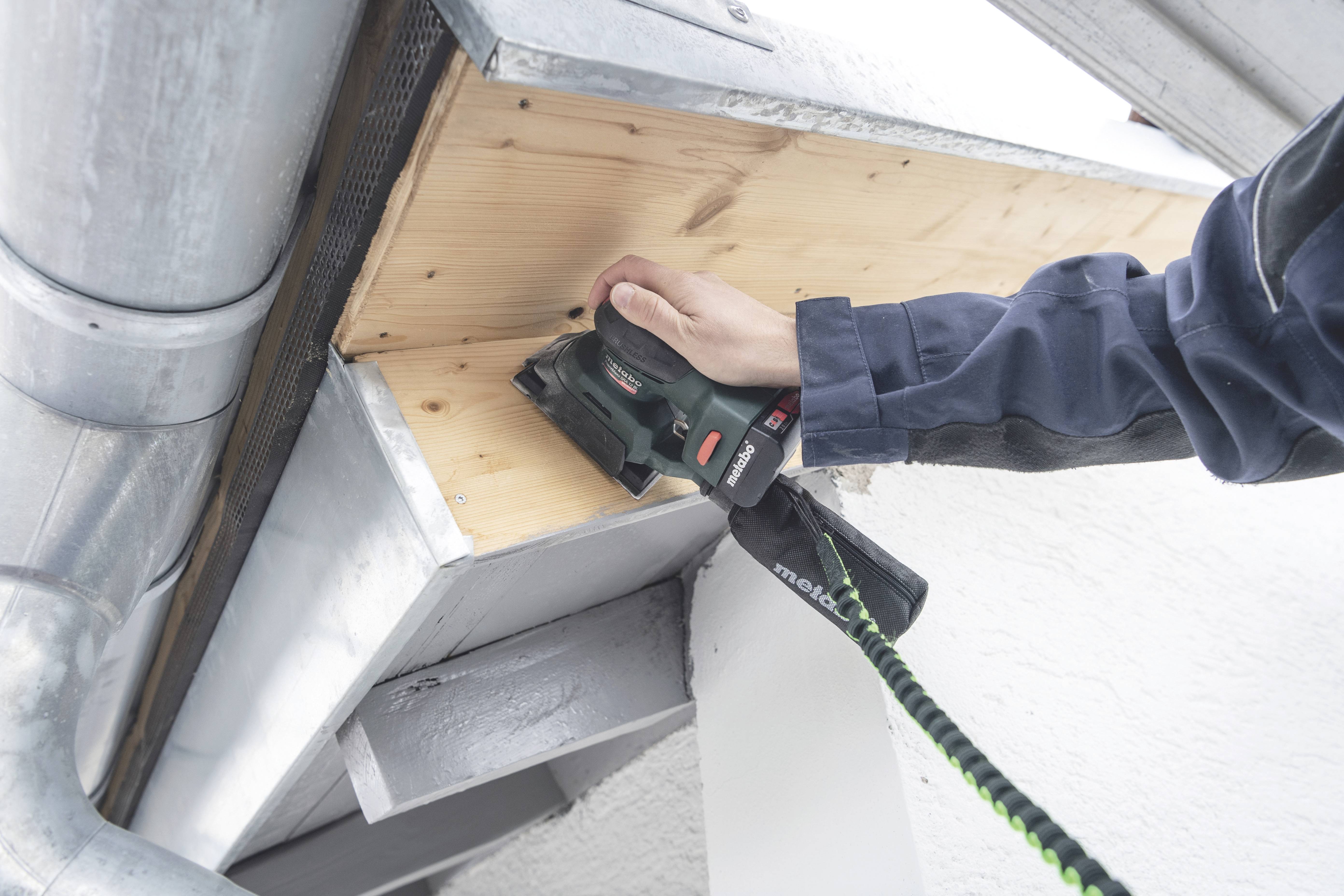 Eine Person schleift Holz an einem Dachvorsprung mit einem Elektroschleifer. Metallteile und Abflussrohre sind sichtbar.