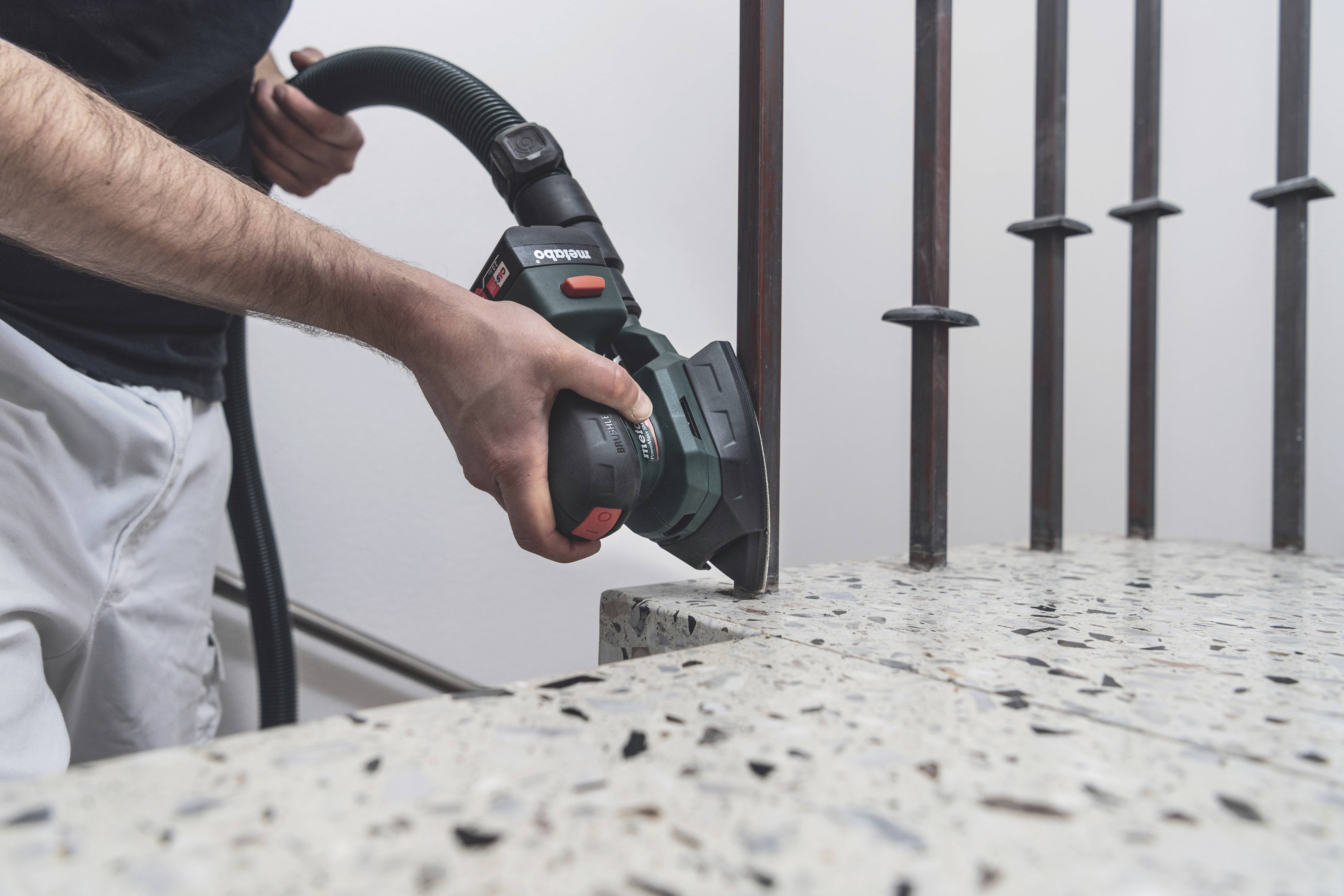 Eine Person schleift mit einem Elektrowerkzeug die Kante eines Beton- oder Steinblocks. Metallstäbe sind im Hintergrund sichtbar.