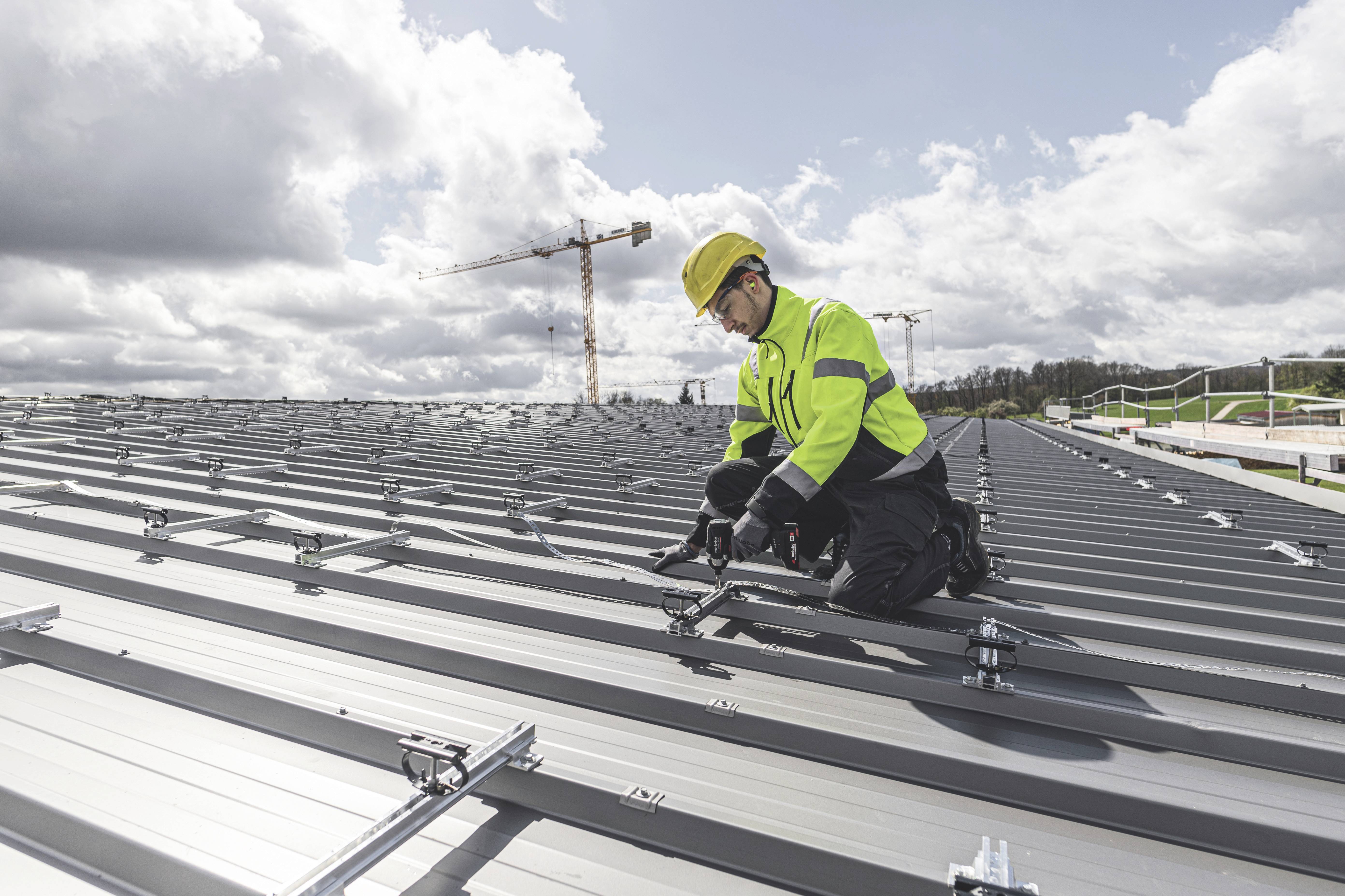 Ein Arbeiter montiert Solarmodule auf einem Metalldach, trägt Schutzkleidung, Helm und Handschuhe. Im Hintergrund ein Baukran und Wolken.