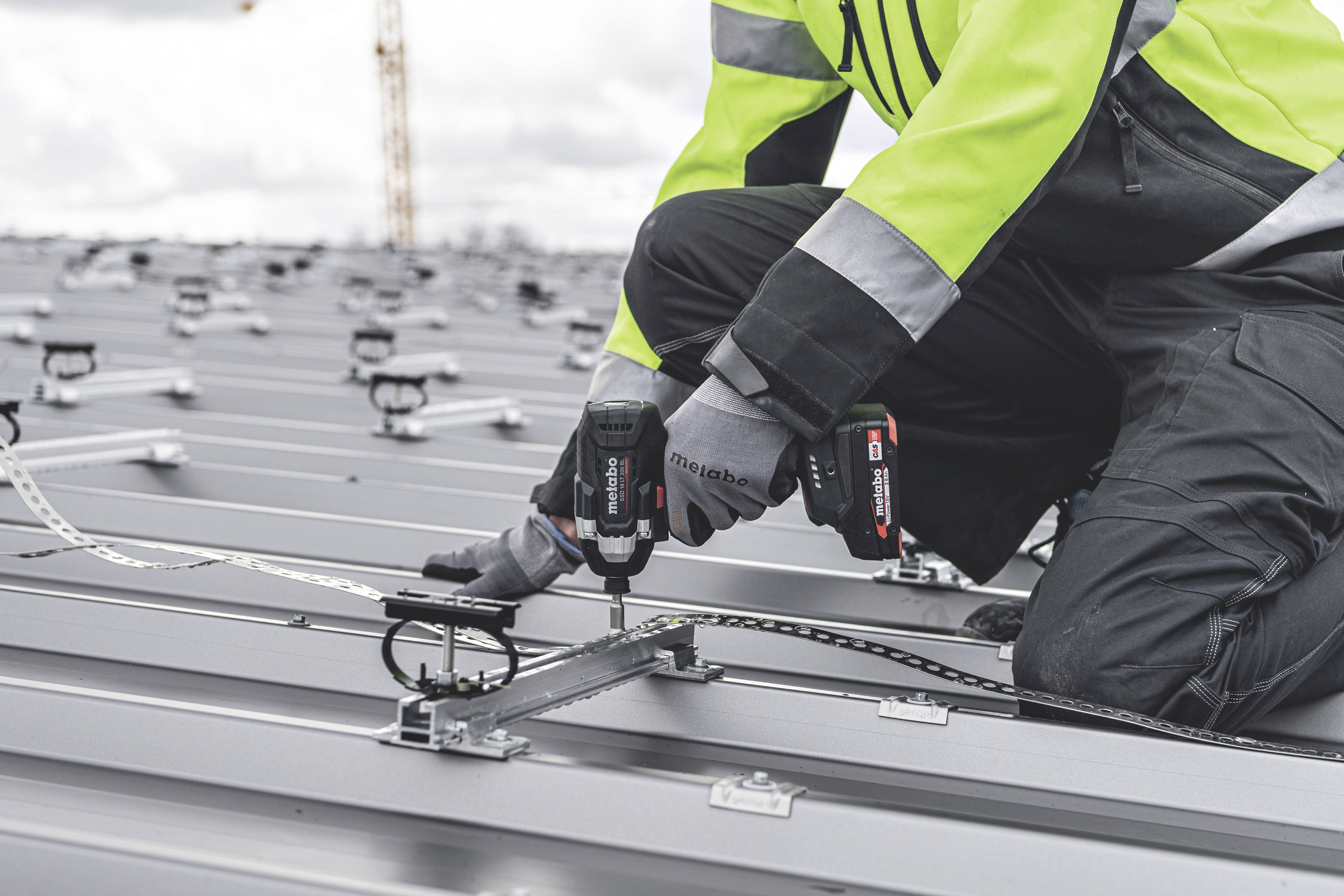 Eine Person in Arbeitskleidung installiert Solarmodule auf einem Metalldach und benutzt dabei eine elektrische Bohrmaschine.