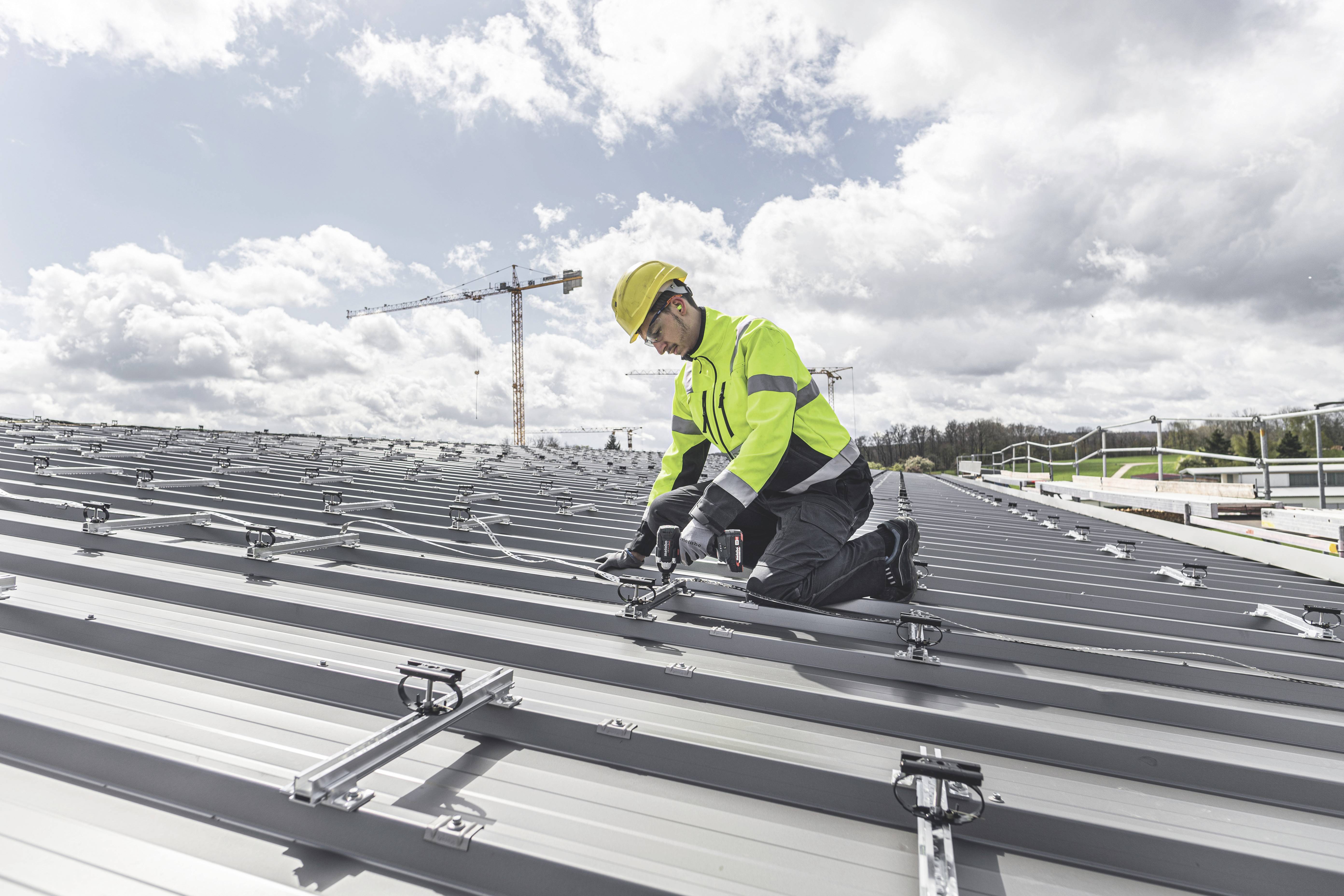 Eine Person in Sicherheitskleidung installiert Solarmodule auf einem großen, silbernen Metalldach. Ein Kran ist im Hintergrund sichtbar.