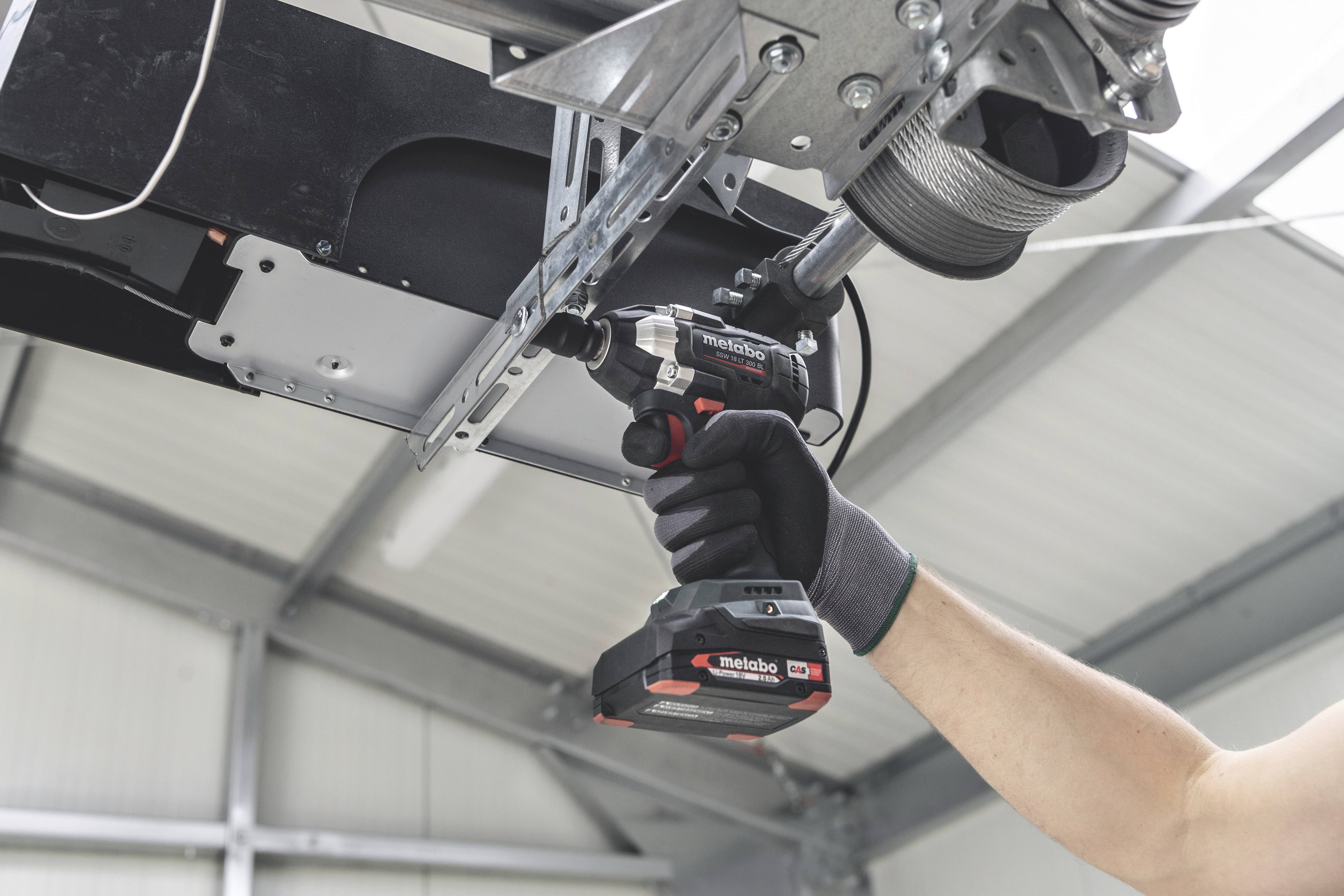 Person montiert mit Akkuschrauber eine Metallvorrichtung an die Decke einer Werkstatt. Sie trägt schwarze Handschuhe.