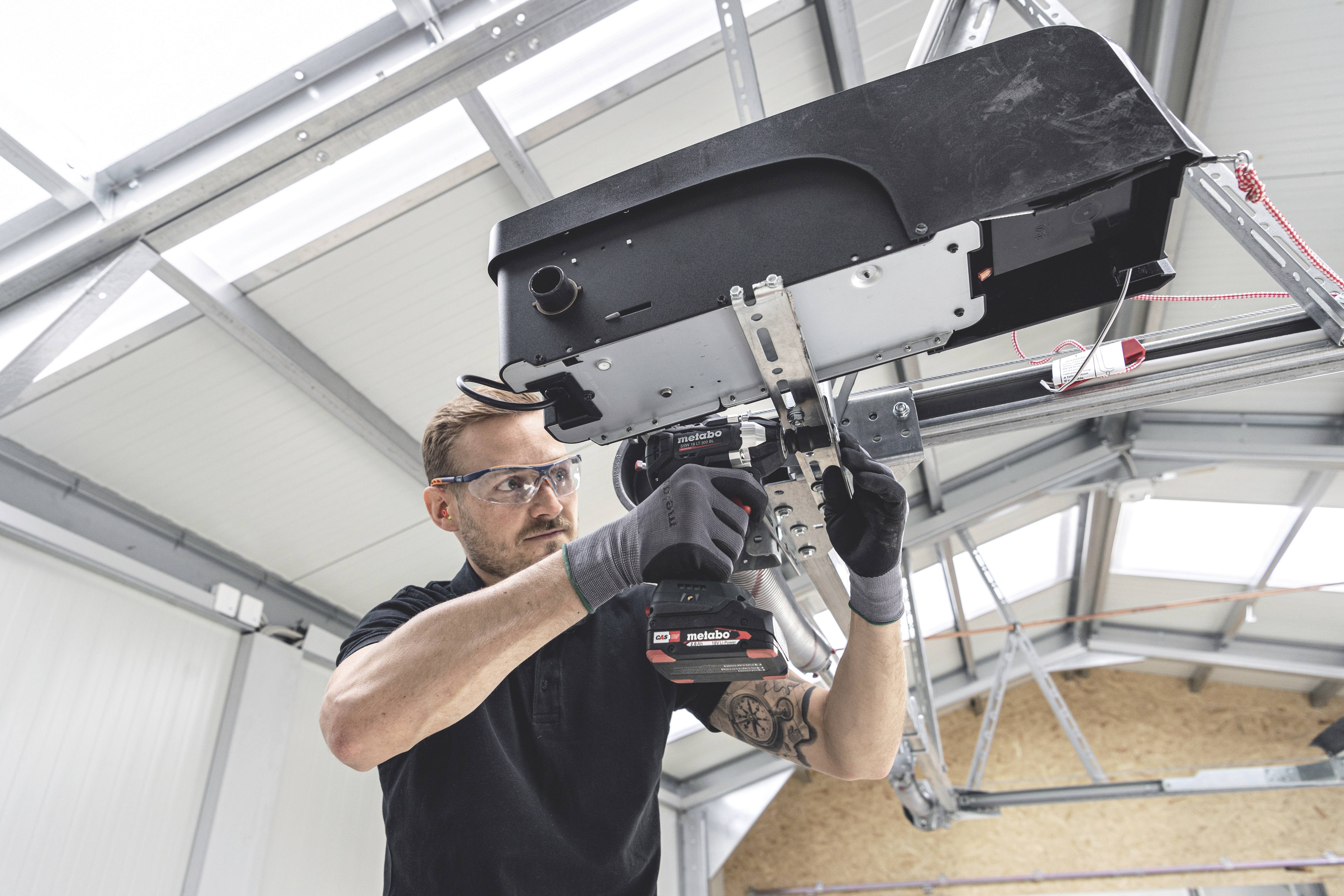 Ein Mann montiert mit einem Akku-Schrauber eine elektrische Torantriebseinheit in einer Werkstatt. Er trägt Schutzbrille und Handschuhe.
