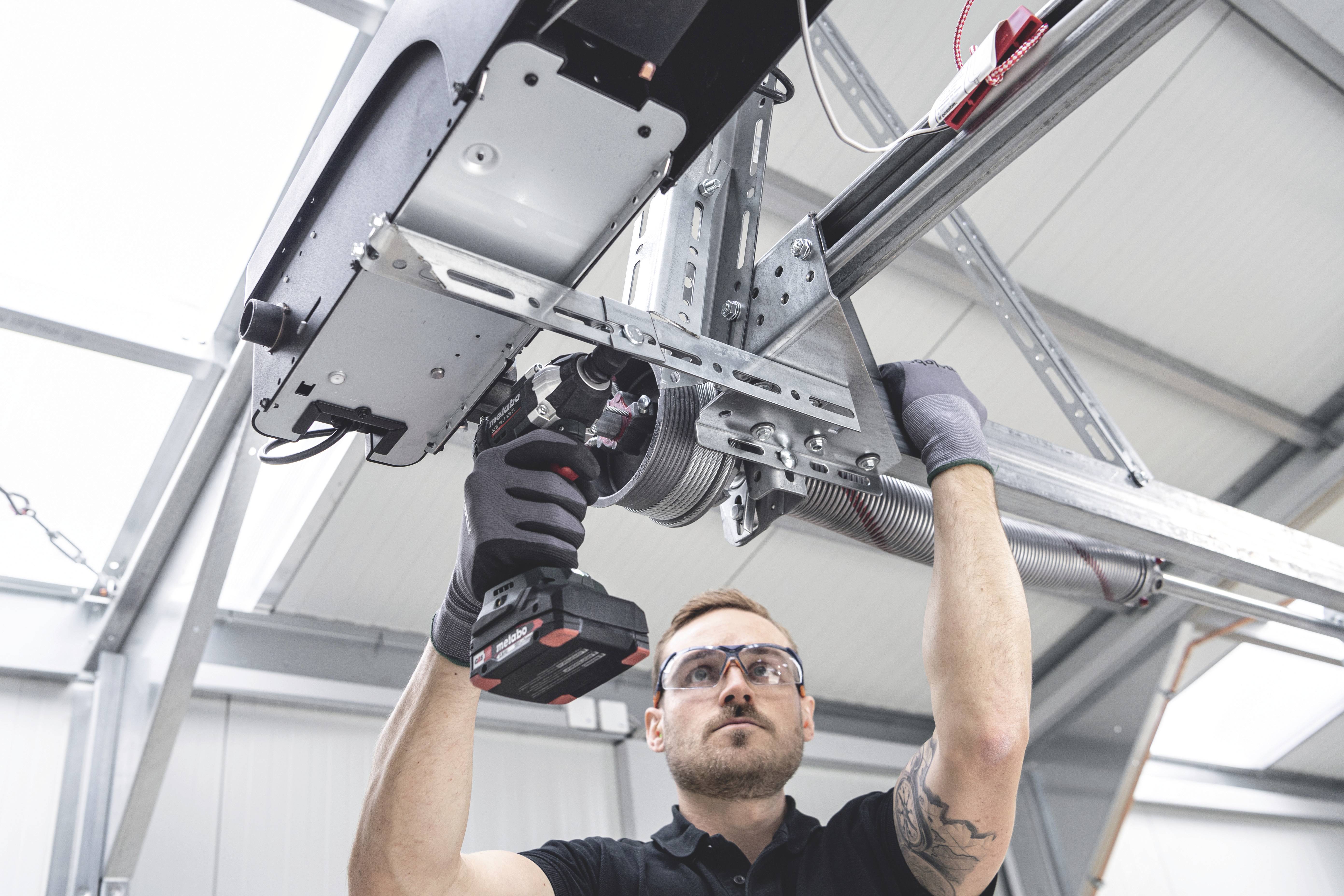 Mann montiert mithilfe eines Akkuschraubers den Motor eines Garagentors. Er trägt Schutzbrille und Arbeitshandschuhe.