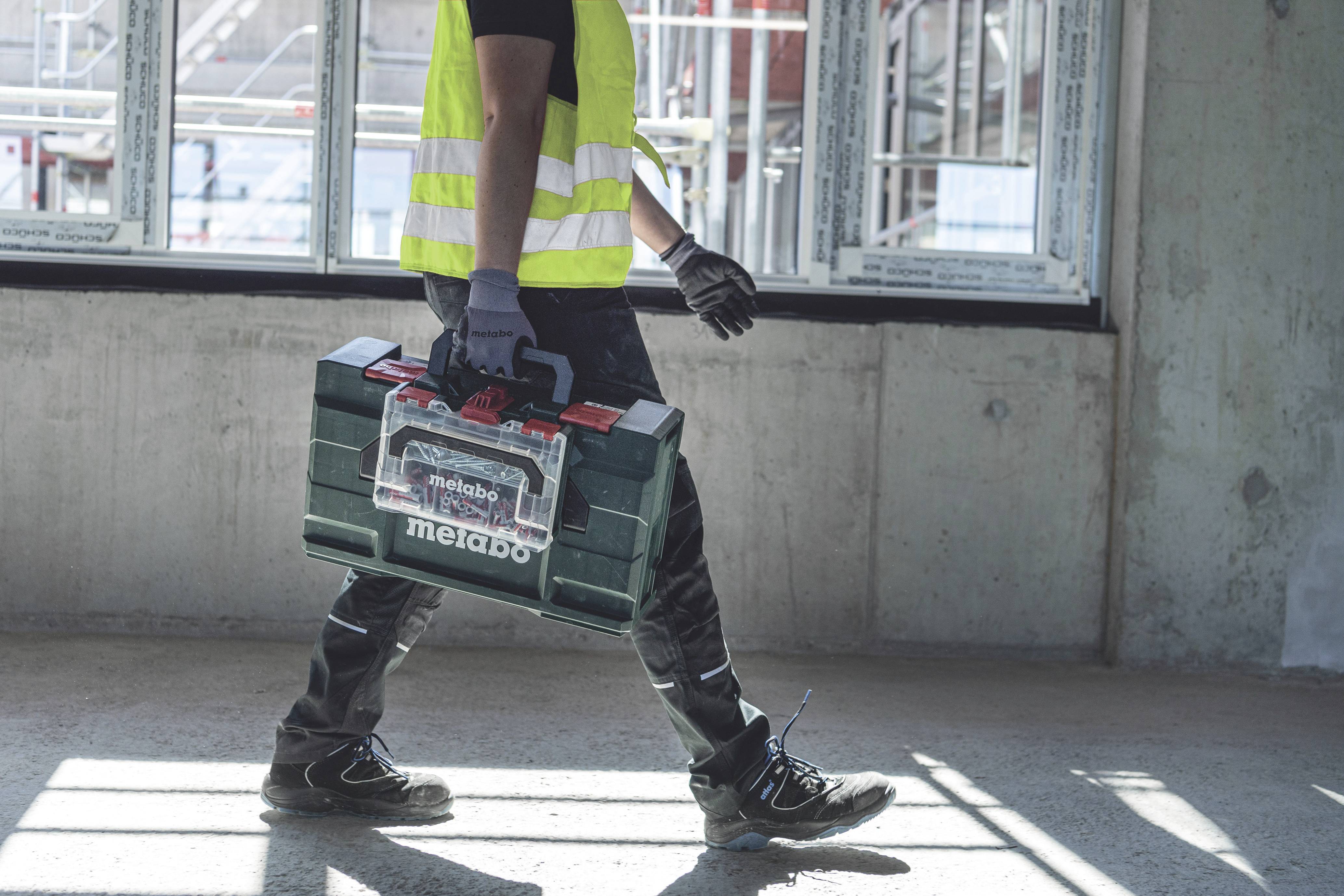 Ein Bauarbeiter in Sicherheitskleidung trägt einen Werkzeugkoffer durch eine Baustelle mit großen Fenstern im Hintergrund.