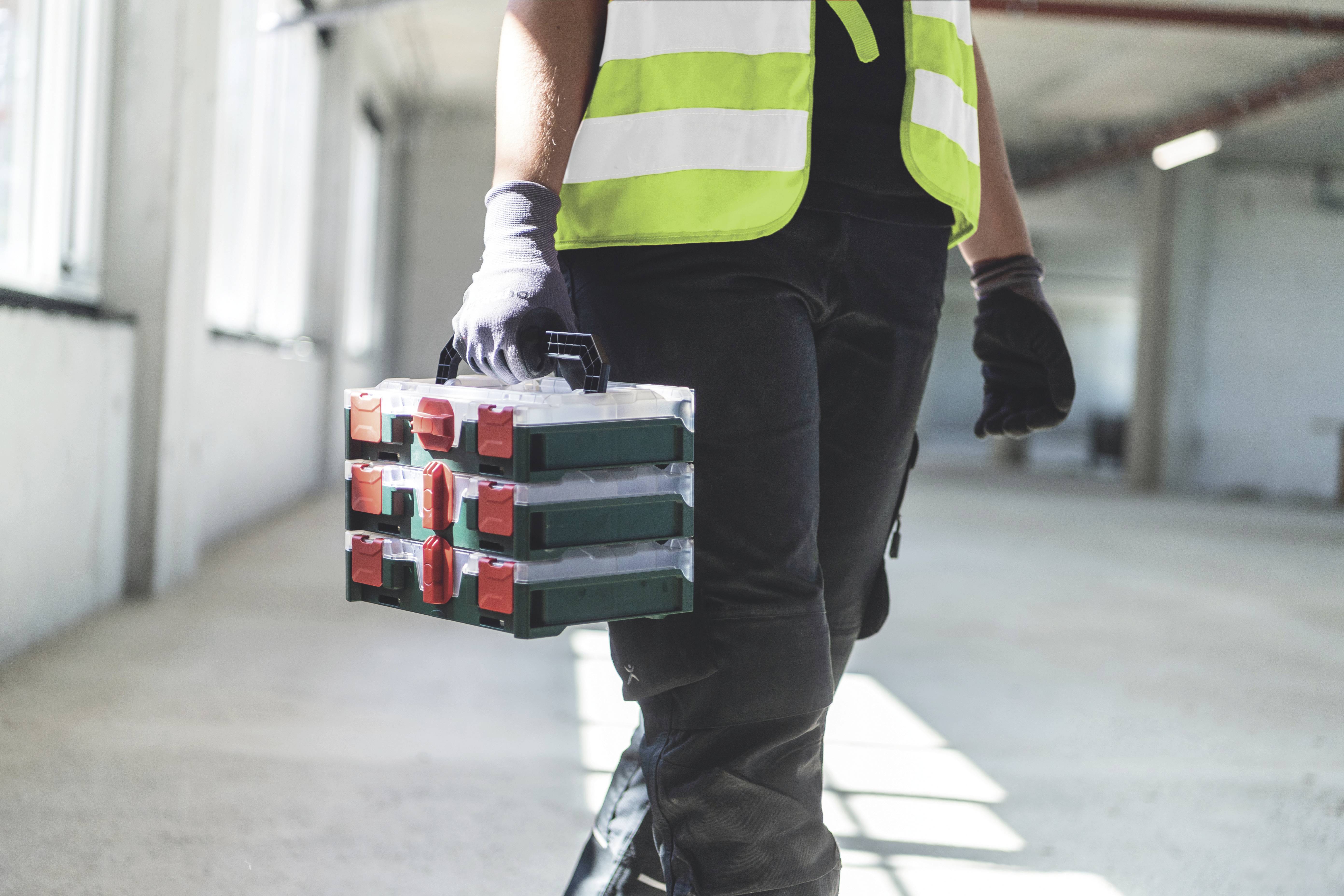 Eine Person in Arbeitskleidung und Sicherheitsweste trägt einen Werkzeugkoffer. Sie befindet sich in einem leeren, hellen Raum.