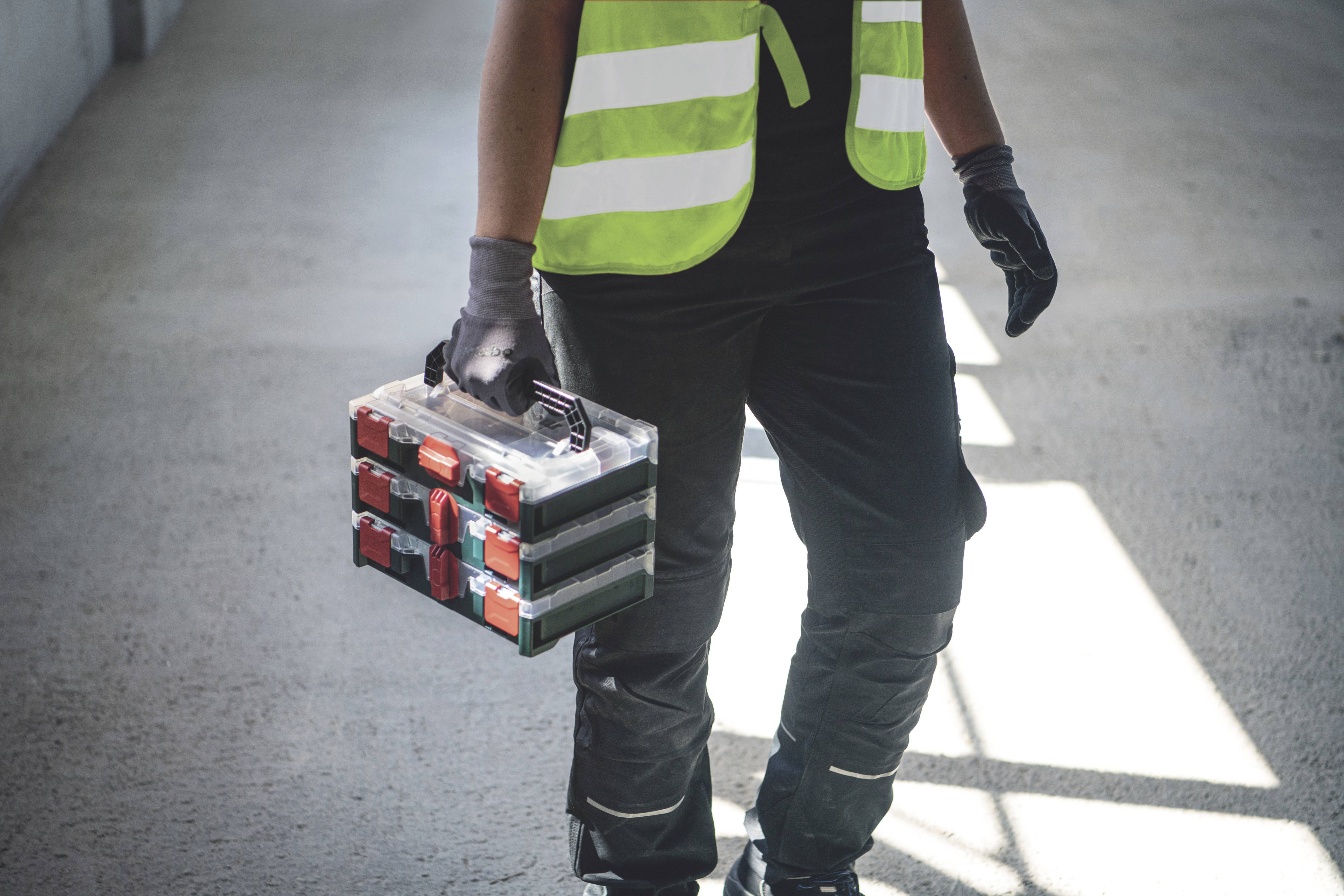 Eine Person in Arbeitskleidung trägt einen Werkzeugkasten in einer lichtdurchfluteten Umgebung.