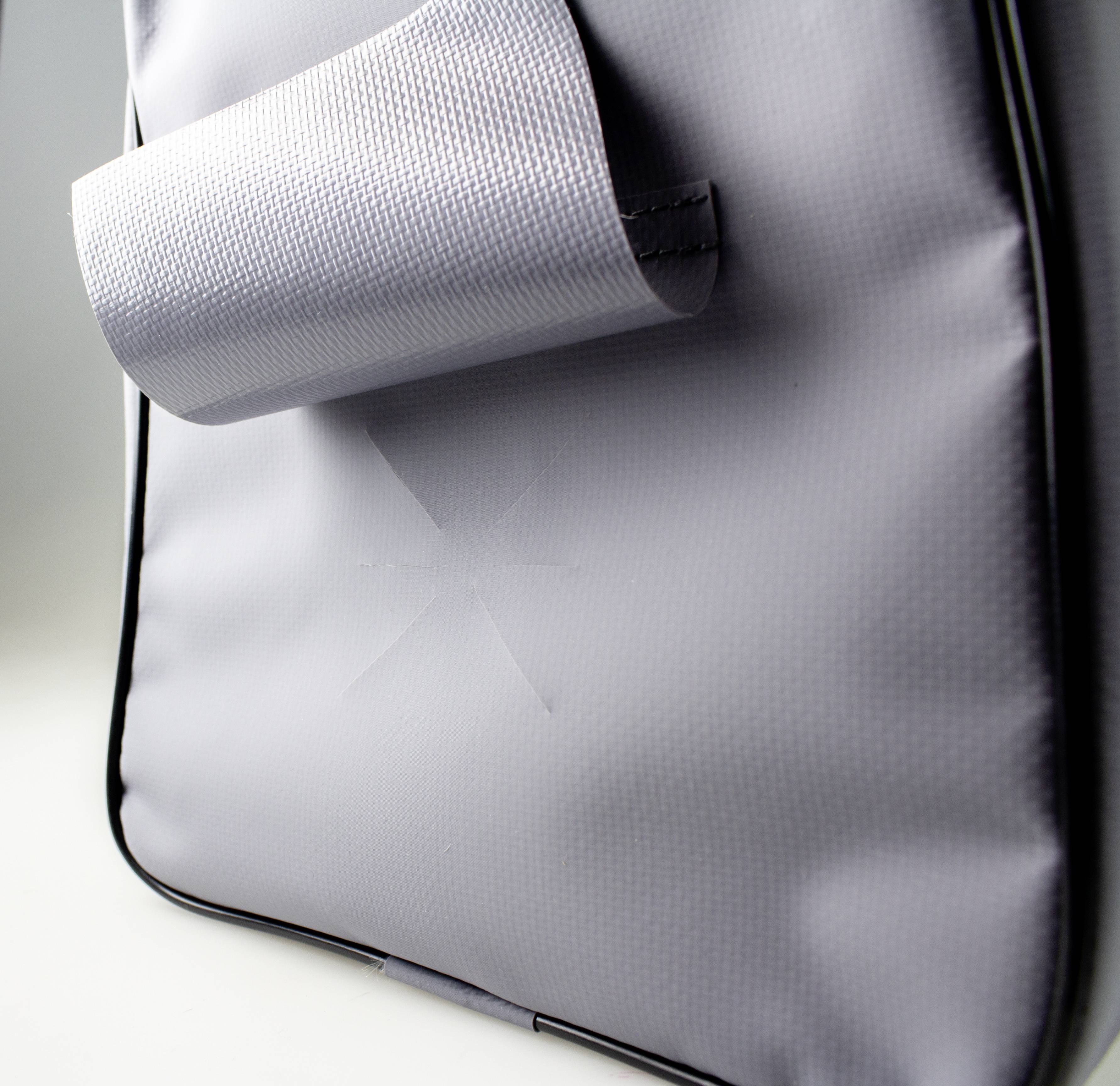 Graues Material einer Tasche mit dünner Struktur. Ein Laschenverschluss ist sichtbar. Die Oberfläche wirkt wasserabweisend.
