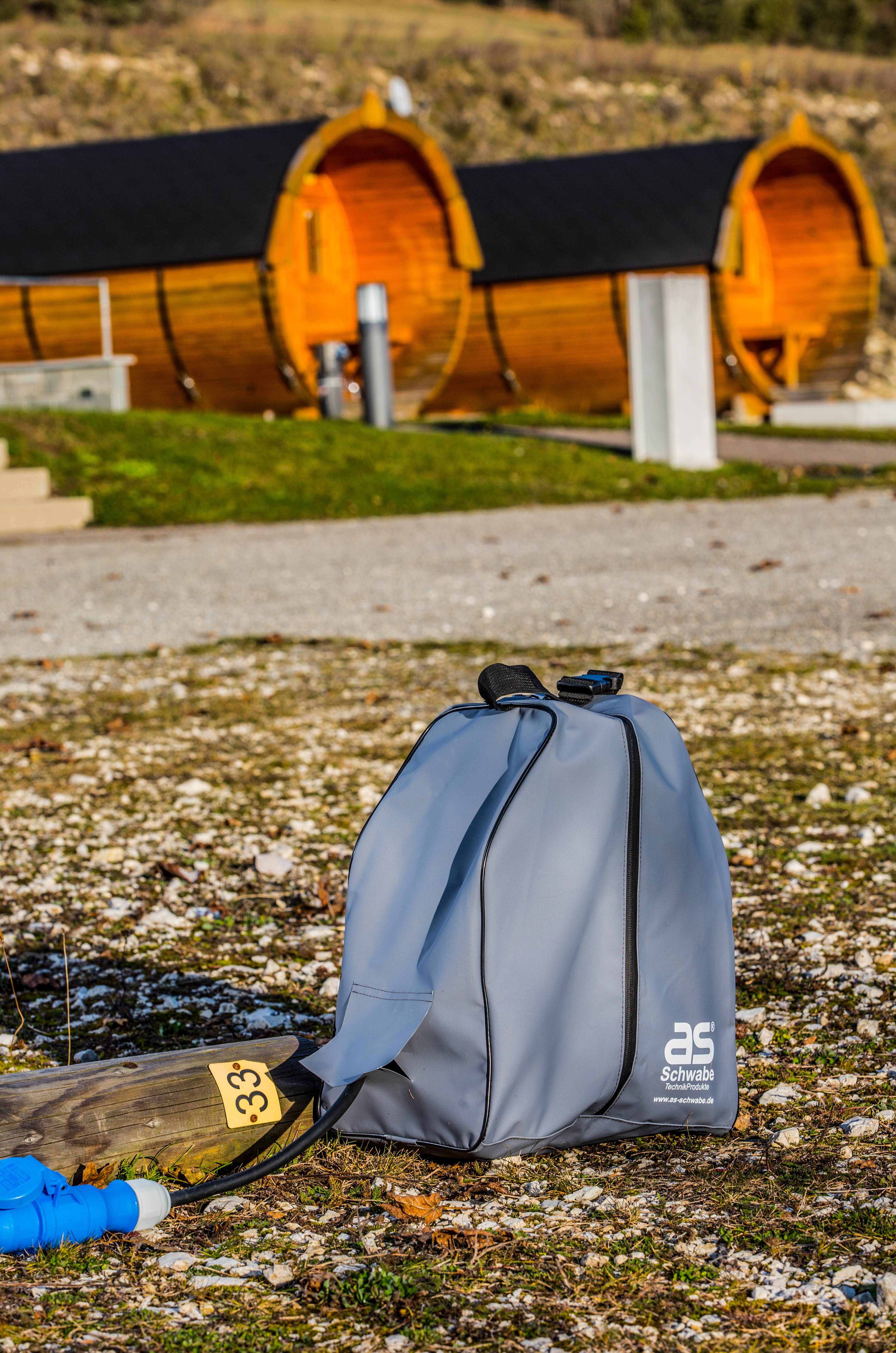 Eine graue Sporttasche steht auf einem Kiesweg im Vordergrund. Im Hintergrund sind Holzhütten mit gewölbten Dächern auf einer Wiese zu sehen.