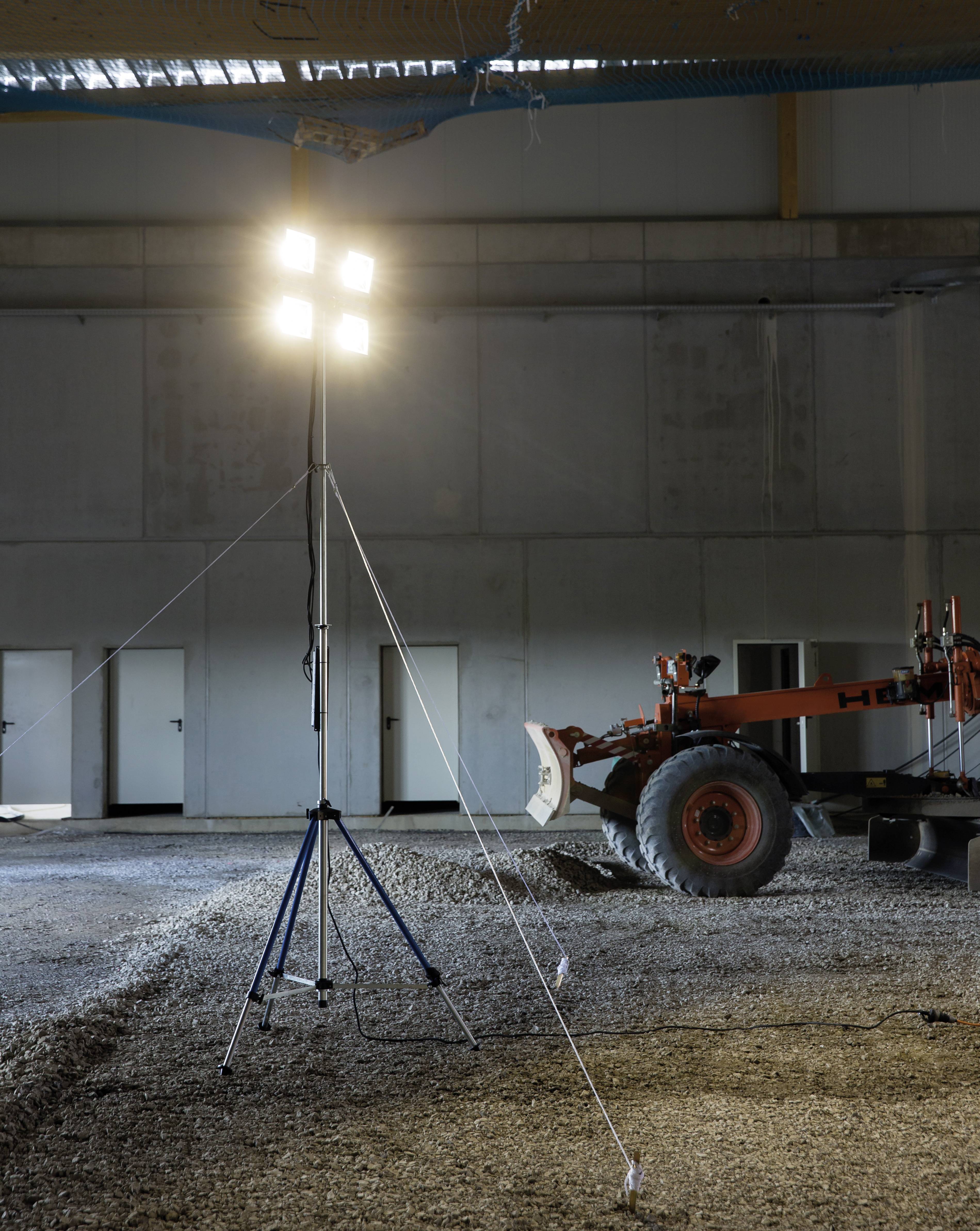 Ein Baustellenstrahler steht in einer teilweise beleuchteten Halle. Im Hintergrund ist ein Baufahrzeug sichtbar.