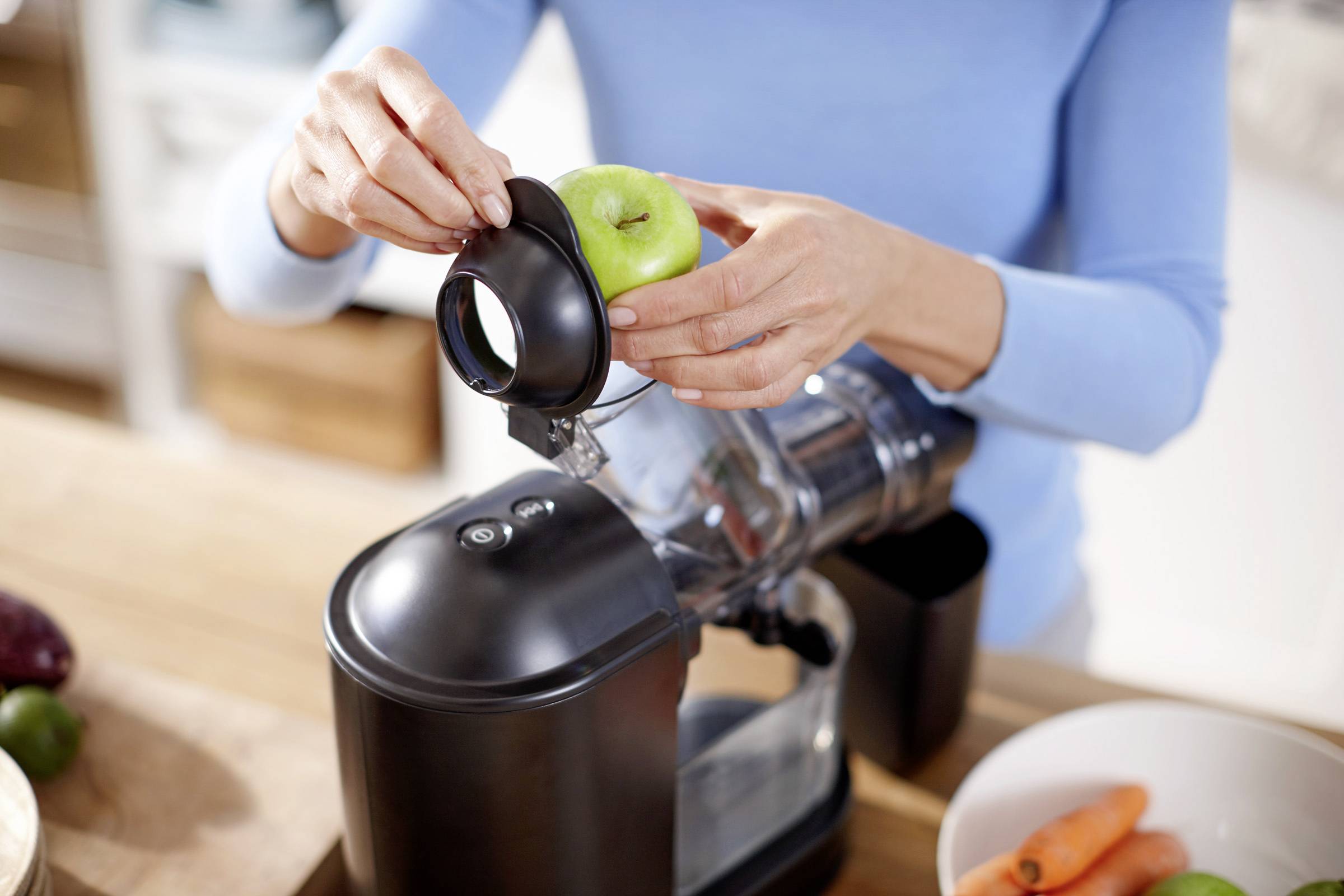 Eine Person steckt einen grünen Apfel in einen schwarzen Entsafter in einer Küche. Daneben liegen Karotten und weiteres Obst.