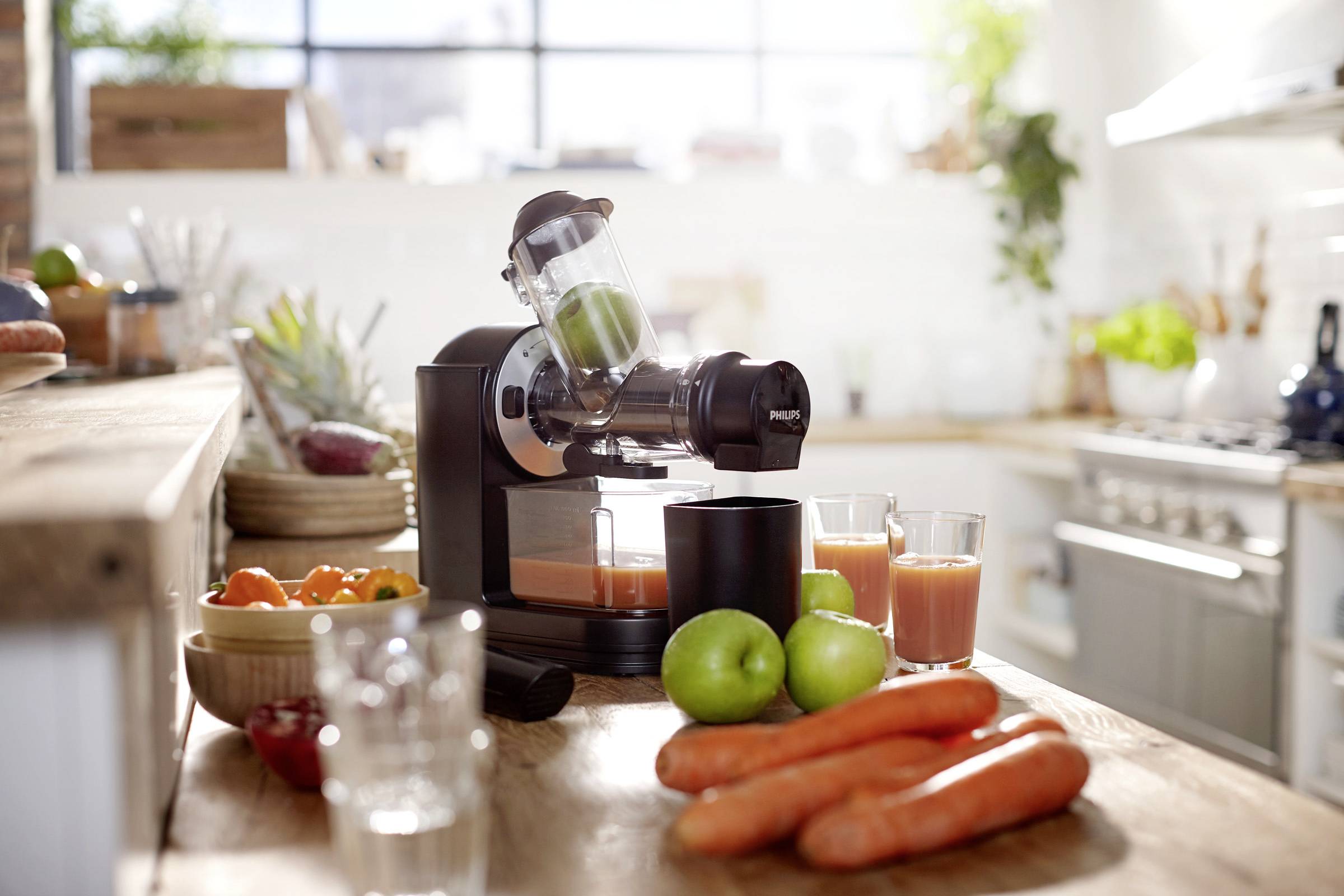 Entsafter auf Küchentheke mit frischem Karottensaft und Gemüse. Helle Küche im Hintergrund, Obst und Gemüse dekorativ arrangiert.