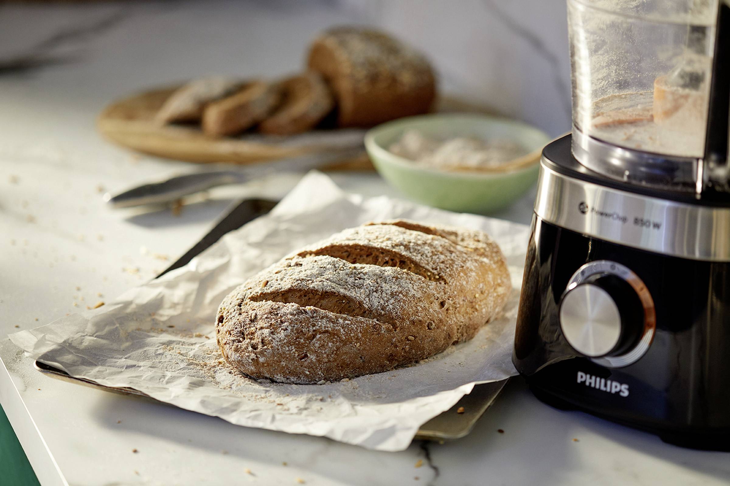 Ein frisch gebackenes Brot liegt auf Backpapier auf einem Tisch, neben einem Mixer. Im Hintergrund sind weitere Brotscheiben.