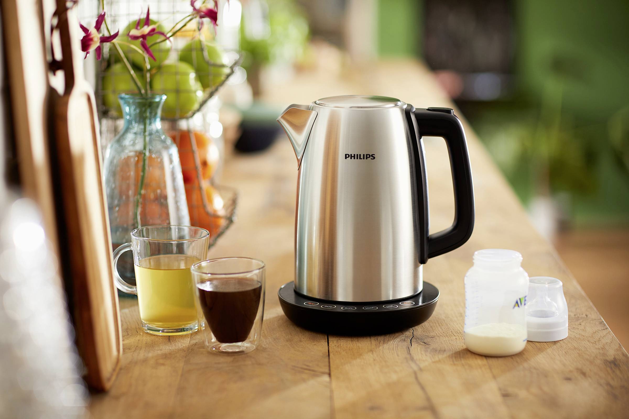 Ein elektrischer Wasserkocher auf einem Holztisch neben einem Glas Saft, einer Tasse Kaffee und einer Milchflasche für Babys.