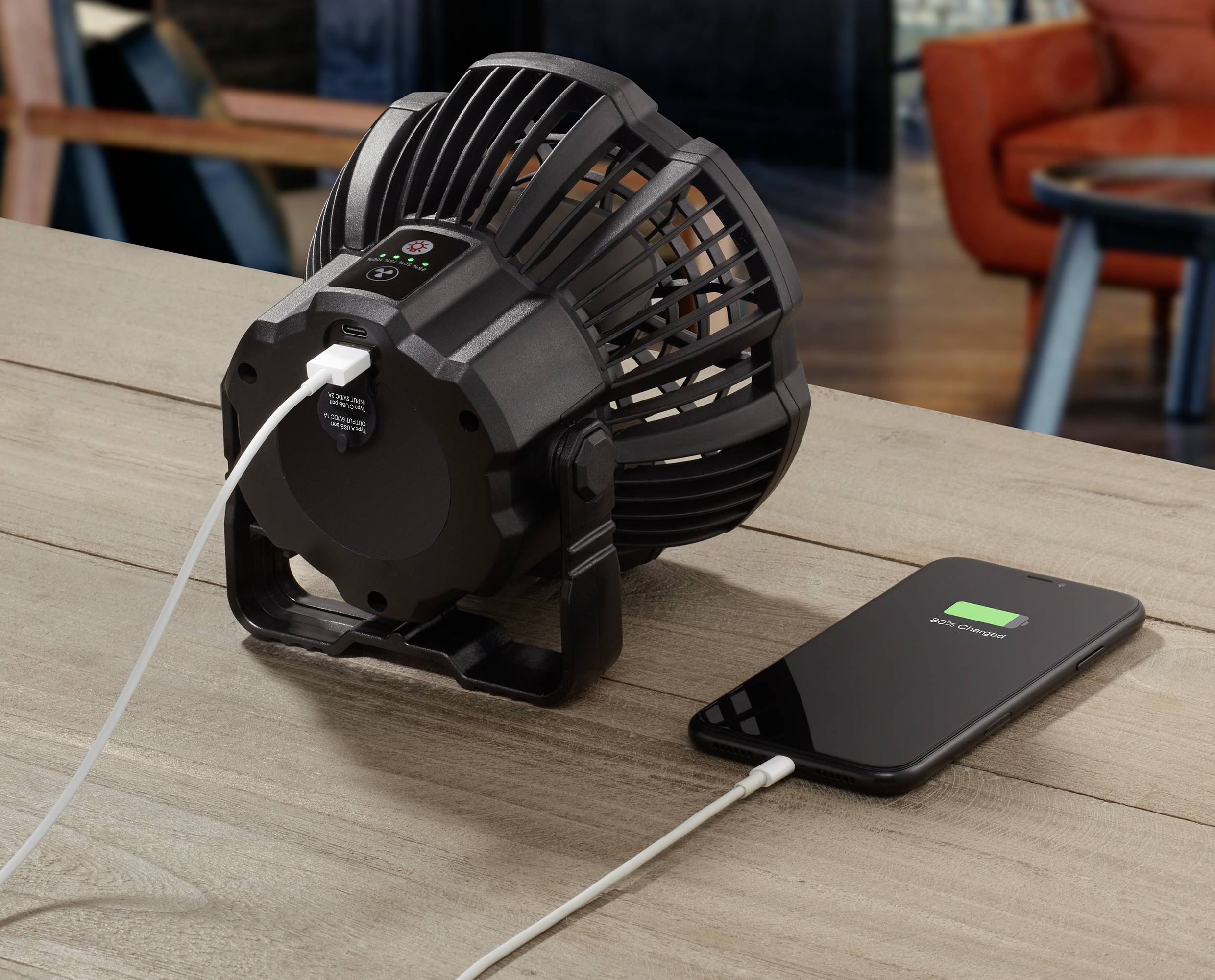 Kleiner Ventilator mit USB-Anschluss lädt ein Smartphone auf einem Holztisch. Hintergrund zeigt unscharfe Möbel.