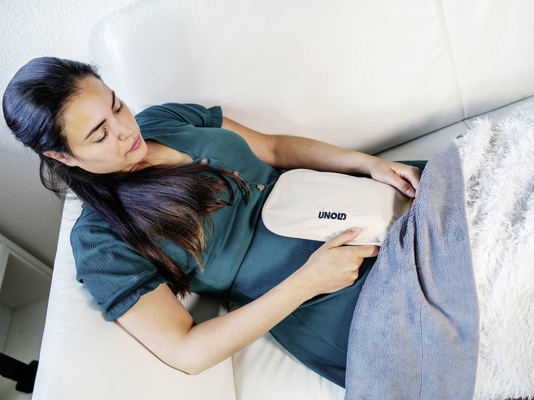 Frau liegt auf einem Sofa, hält ein Heizkissen auf ihrem Bauch. Sie trägt ein grünes Kleid und liegt unter einer grauen Decke.