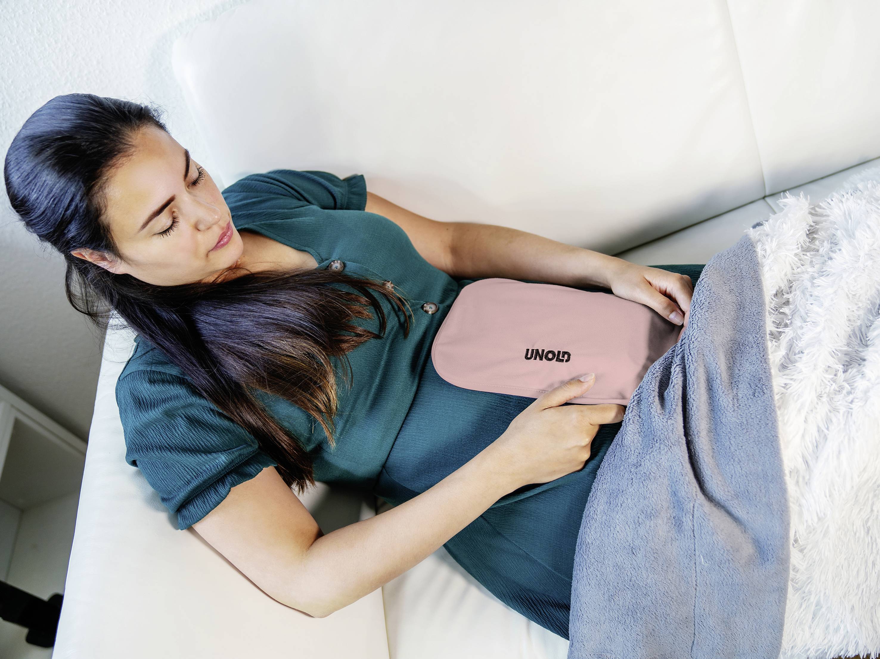 Eine Frau liegt auf einem Sofa und hält ein rosafarbenes Wärmekissen auf ihrem Bauch. Sie trägt ein grünes Kleid und entspannt sich.