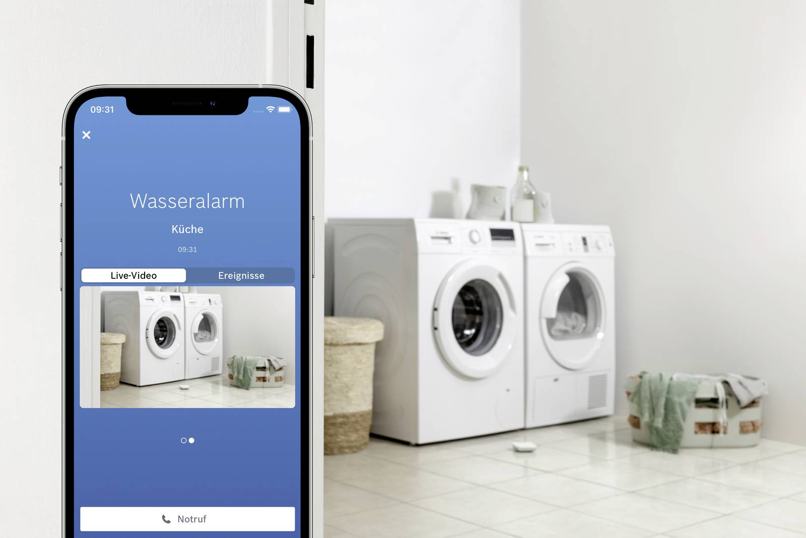 Ein Smartphone zeigt einen 'Wasseralarm' in der Küche an. Im Hintergrund stehen Waschmaschine und Trockner, umgeben von Wäschekörben.