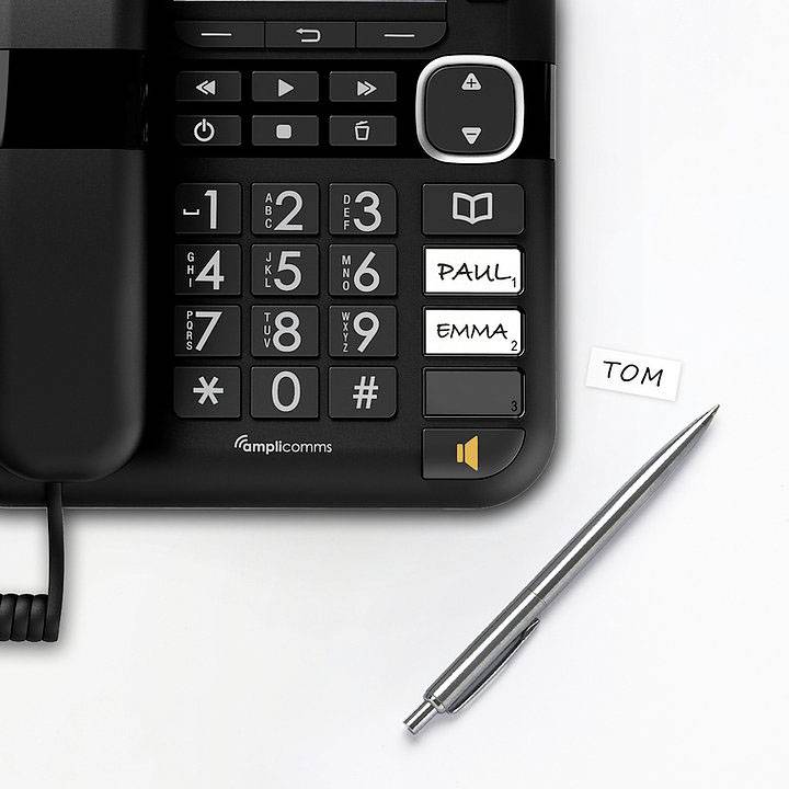 Ein schwarzes Telefon mit großen Tasten, neben einem Stift und einem Zettel mit der Aufschrift 'TOM', auf dem Display steht 'PAUL, EMMA'.