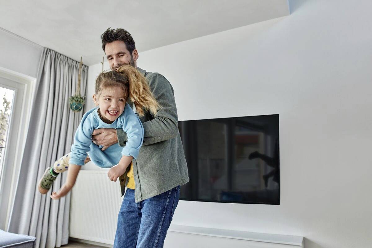 Ein Vater hält sein lachendes Kind spielerisch in der Luft im Wohnzimmer. Ein Fernseher ist an der Wand im Hintergrund sichtbar.