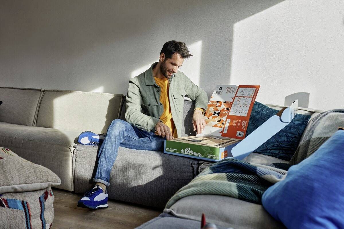 Ein Mann sitzt auf einem Sofa und öffnet eine Essenslieferbox. Er sieht zufrieden aus, während Sonnenlicht durch ein Fenster scheint.