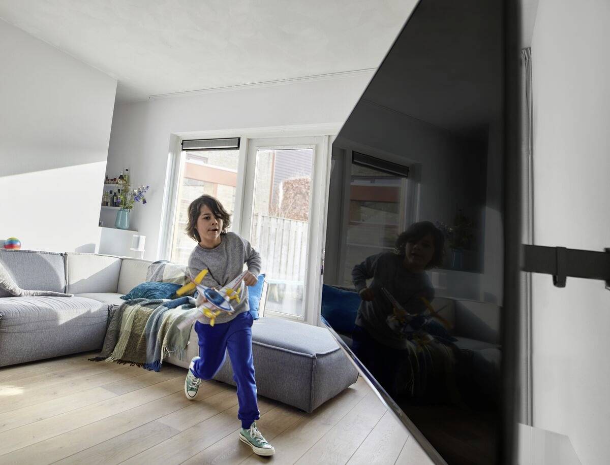 Ein Kind spielt fröhlich im Wohnzimmer mit einem Spielzeugflugzeug. Im Hintergrund ist ein großes Sofa und ein Fenster zu sehen.