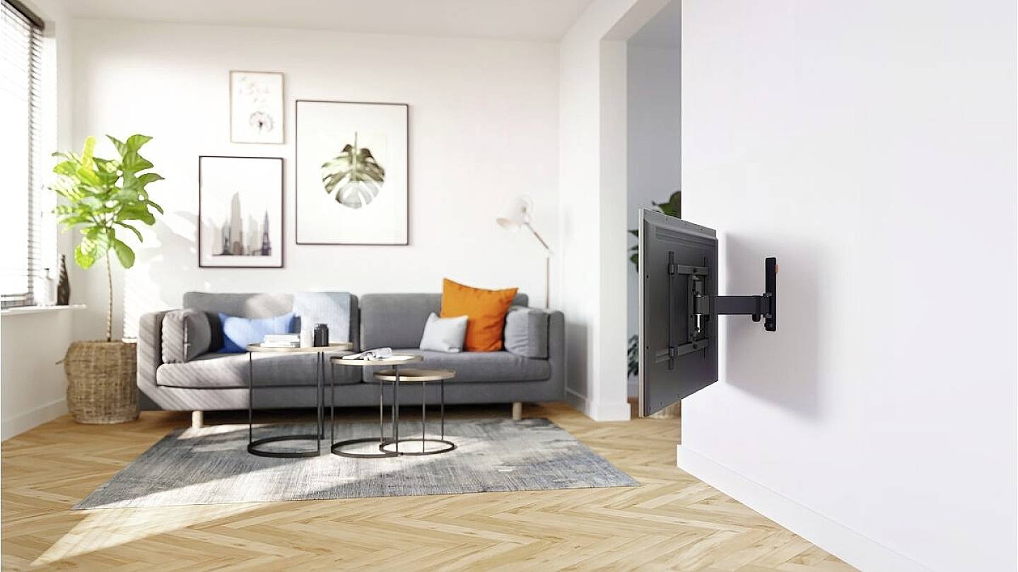 Helles Wohnzimmer mit grauem Sofa, bunte Kissen, Zimmerpflanze, Wandbildern und einem an der Wand montierten Fernseher.