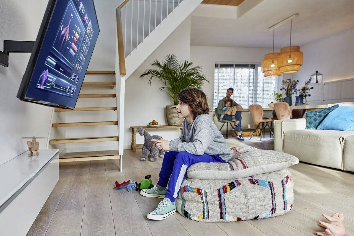 Ein Junge sitzt auf einem Kissen und spielt Videospiele auf einem großen Fernseher. Im Hintergrund sitzen zwei Erwachsene auf einem Sofa.