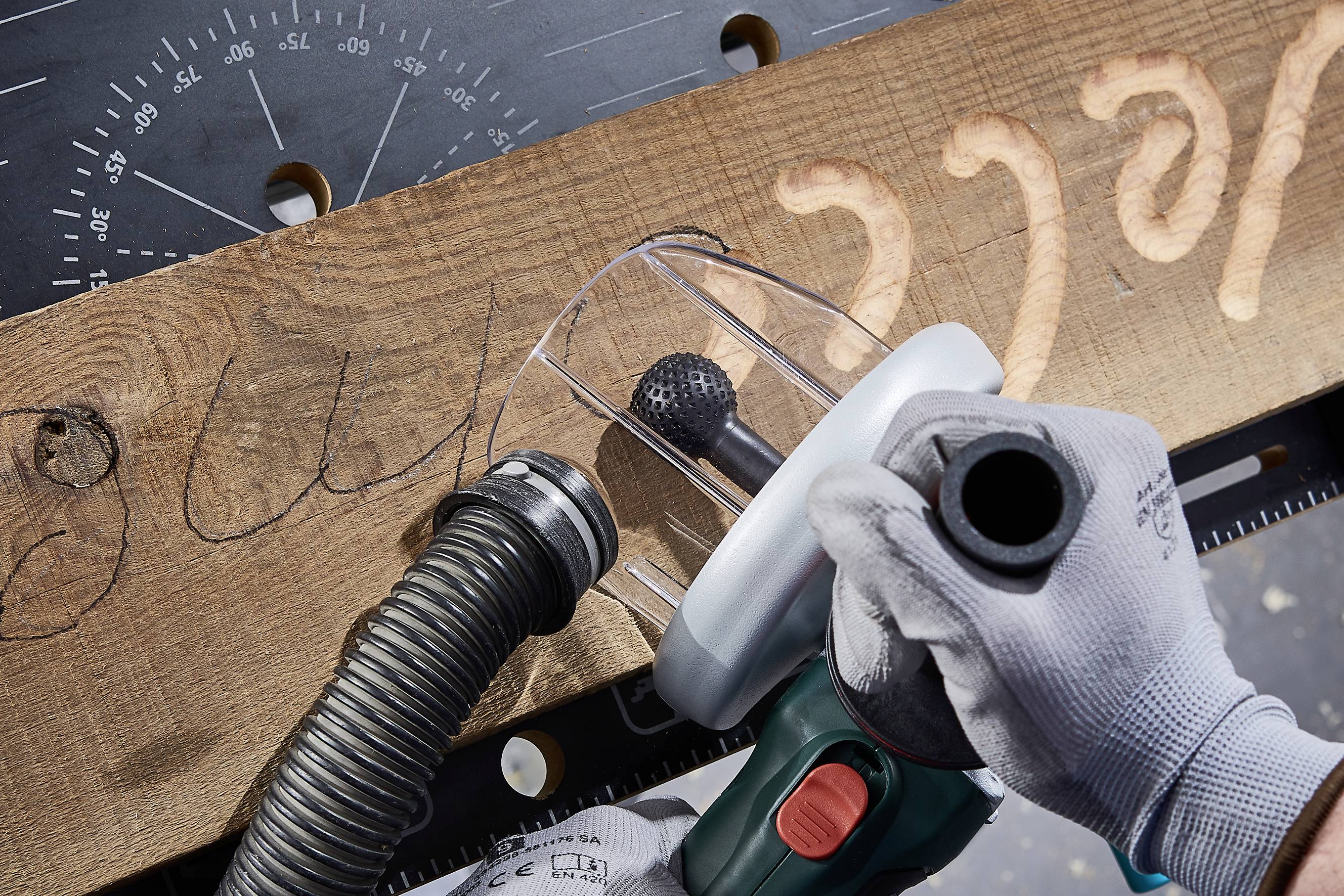 Ein Holzbrett wird mit einem Handwerkzeug bearbeitet. Ein Arbeiter trägt Handschuhe und verwendet eine Absaugvorrichtung, um die Werkstatt sauber zu halten.