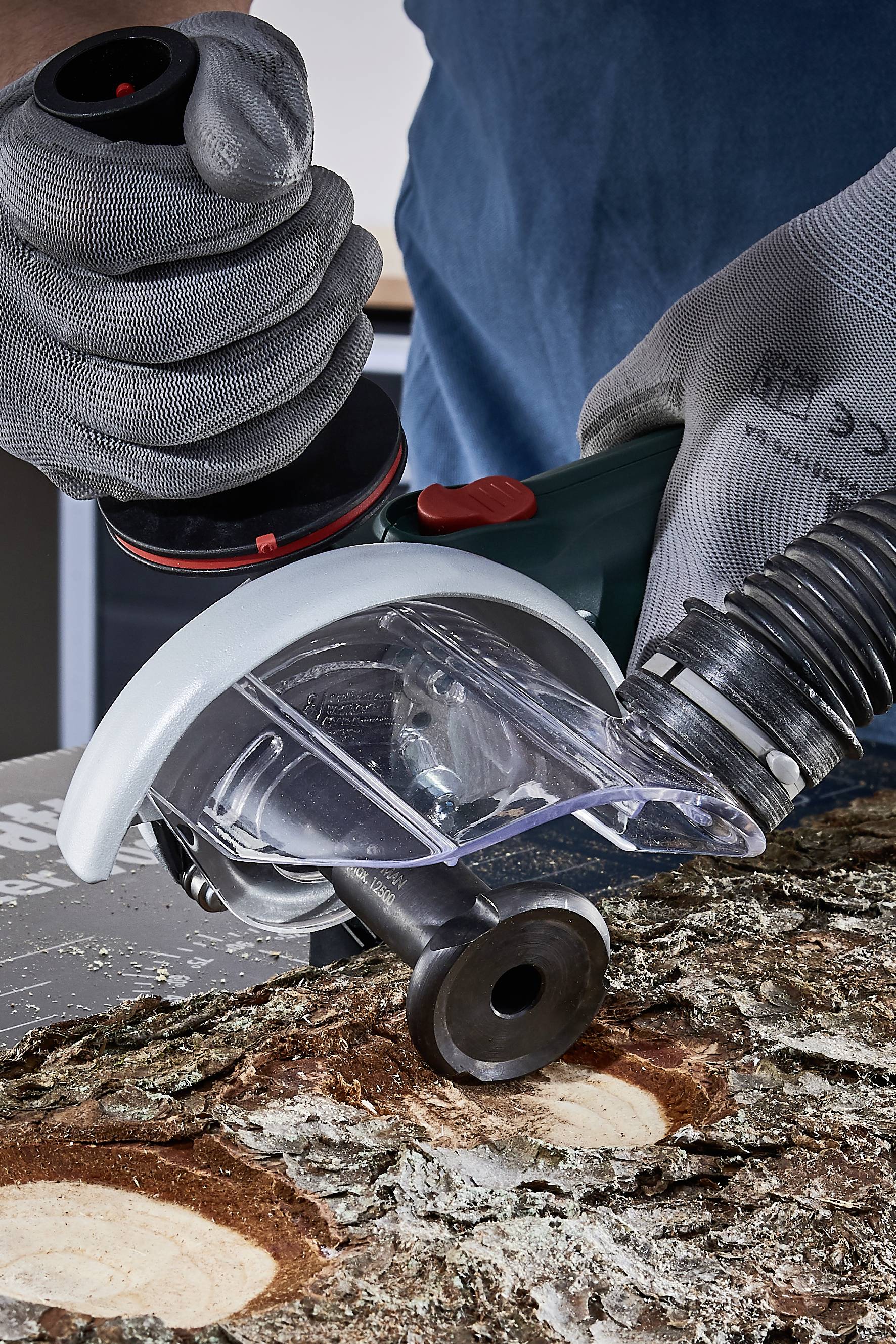 Eine Person trägt Schutzhandschuhe und hält ein Elektrowerkzeug, das Holz bearbeitet. Späne und Baumrinde sind sichtbar.