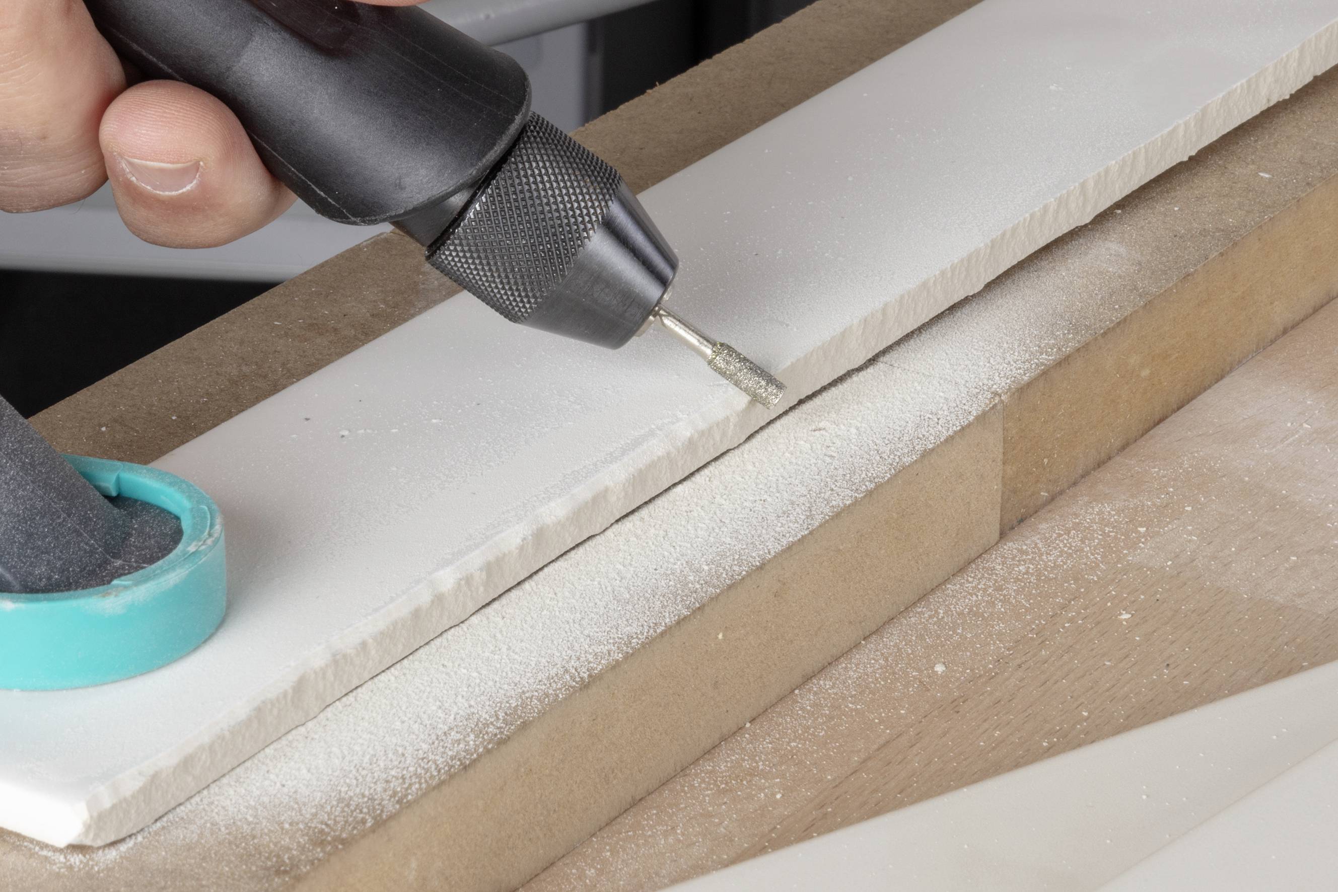 Eine Hand hält ein rotierendes Werkzeug, das eine weiße Fliese auf einem Tisch glättet. Staubpartikel sind sichtbar.