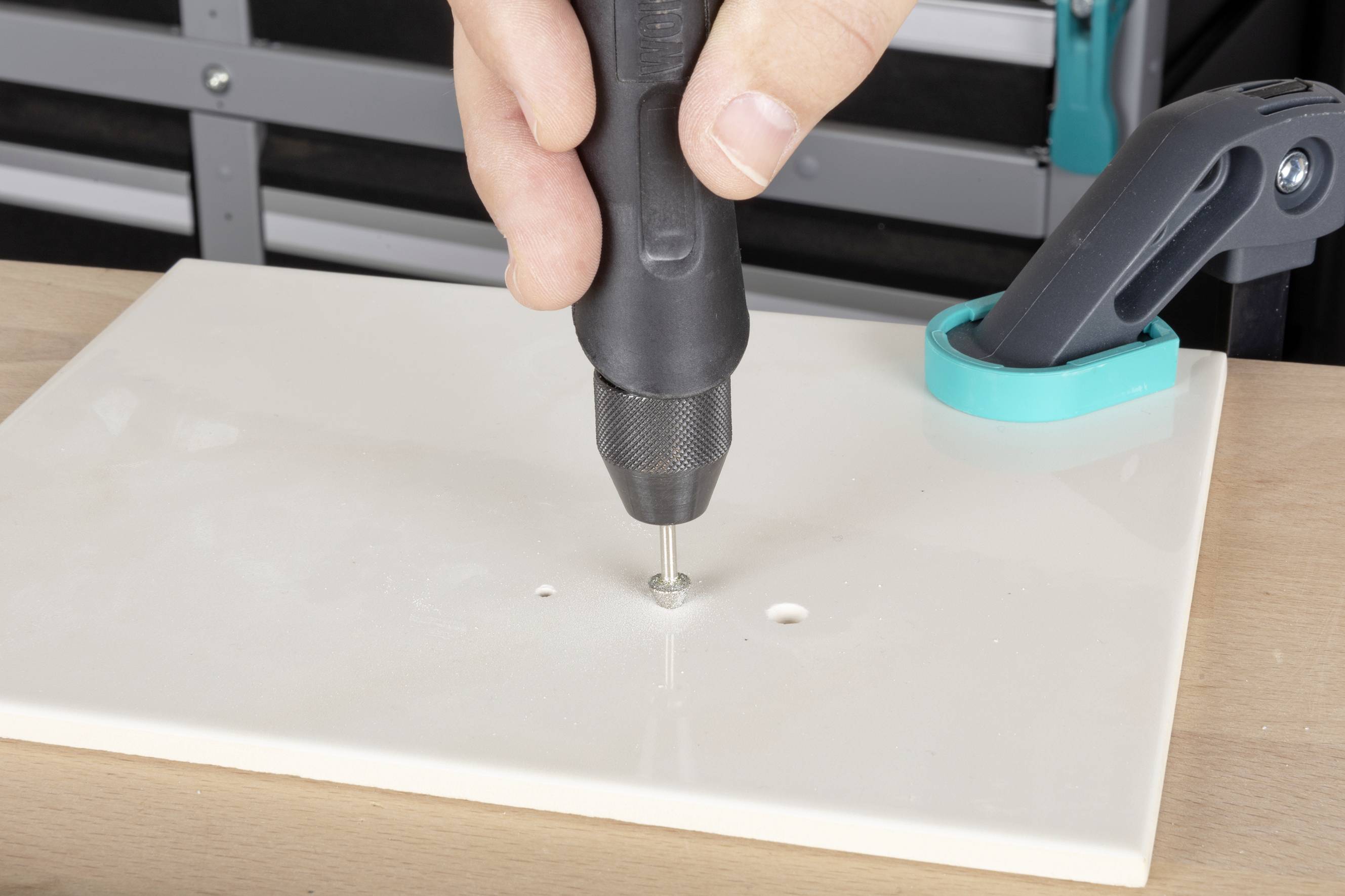 Eine Hand hält einen Bohrer, der ein kleines Loch in eine weiße Platte bohrt, die mit einer blauen Klemme auf einem Tisch befestigt ist.