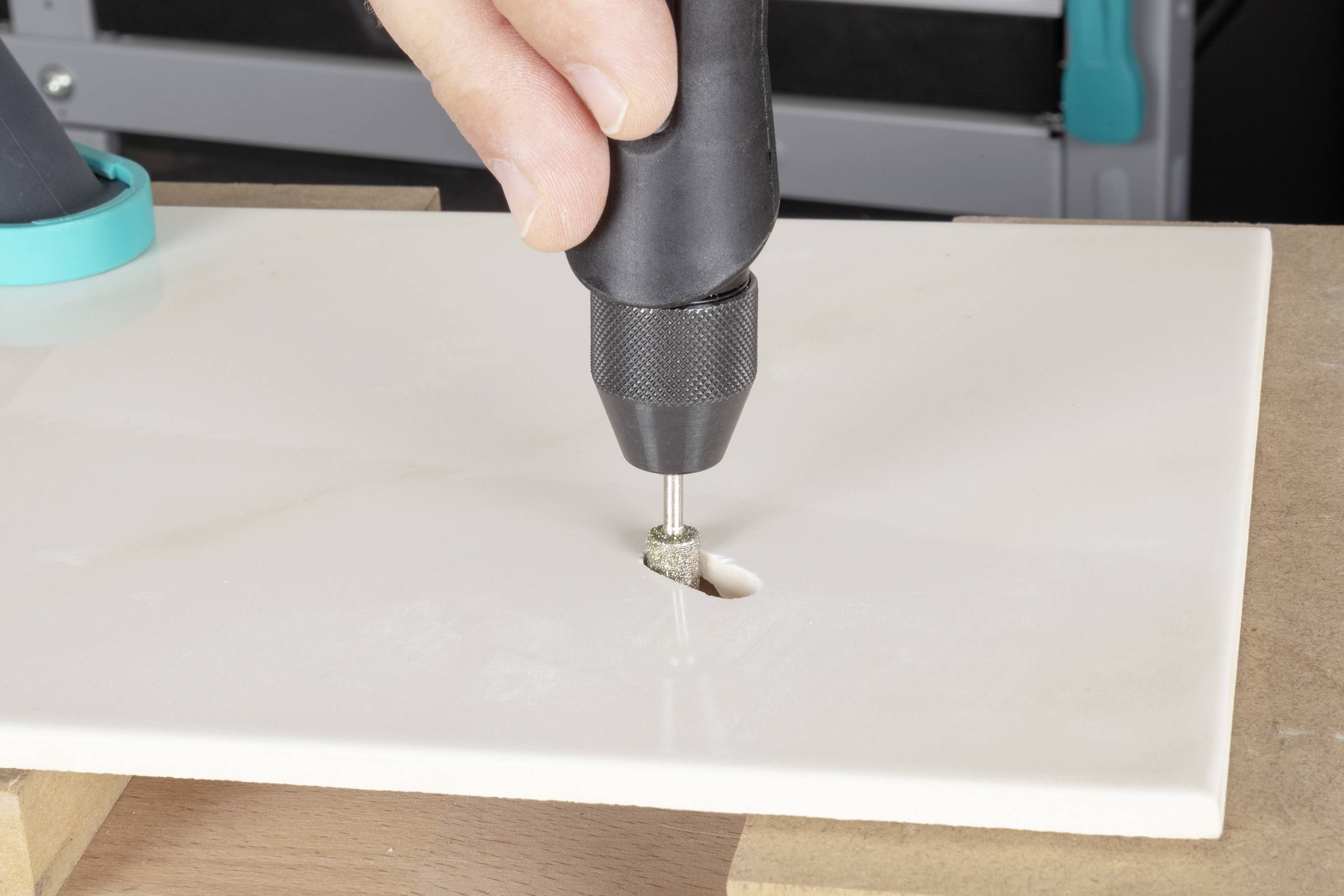 Hand hält einen Bohrer, der ein Loch in eine weiße Keramikfliese bohrt. Fliese liegt auf einem Holzbrett.