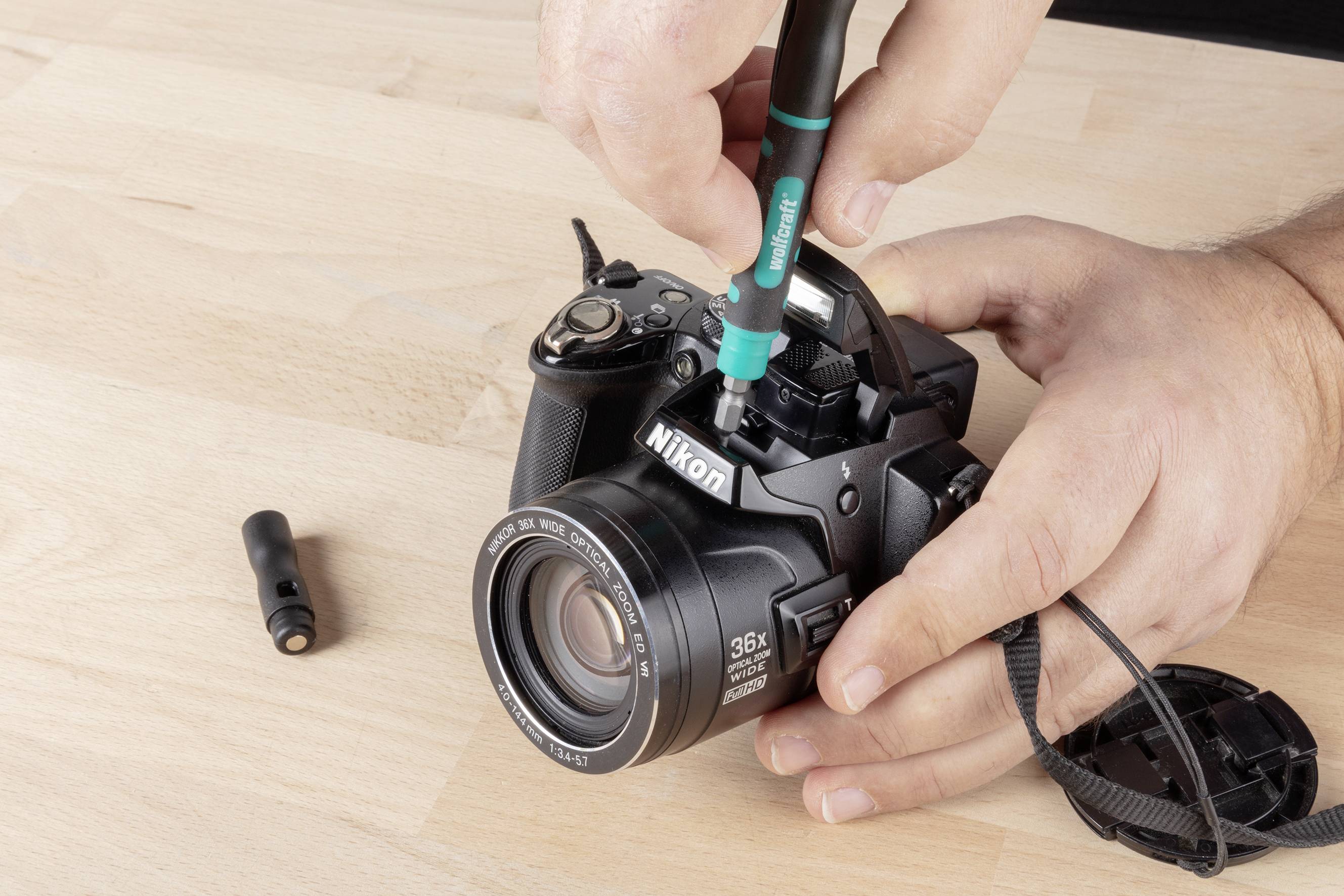 Eine Person reinigt die Linse einer Nikon-Kamera mit einem speziellen Reinigungsstift auf einem Holztisch.