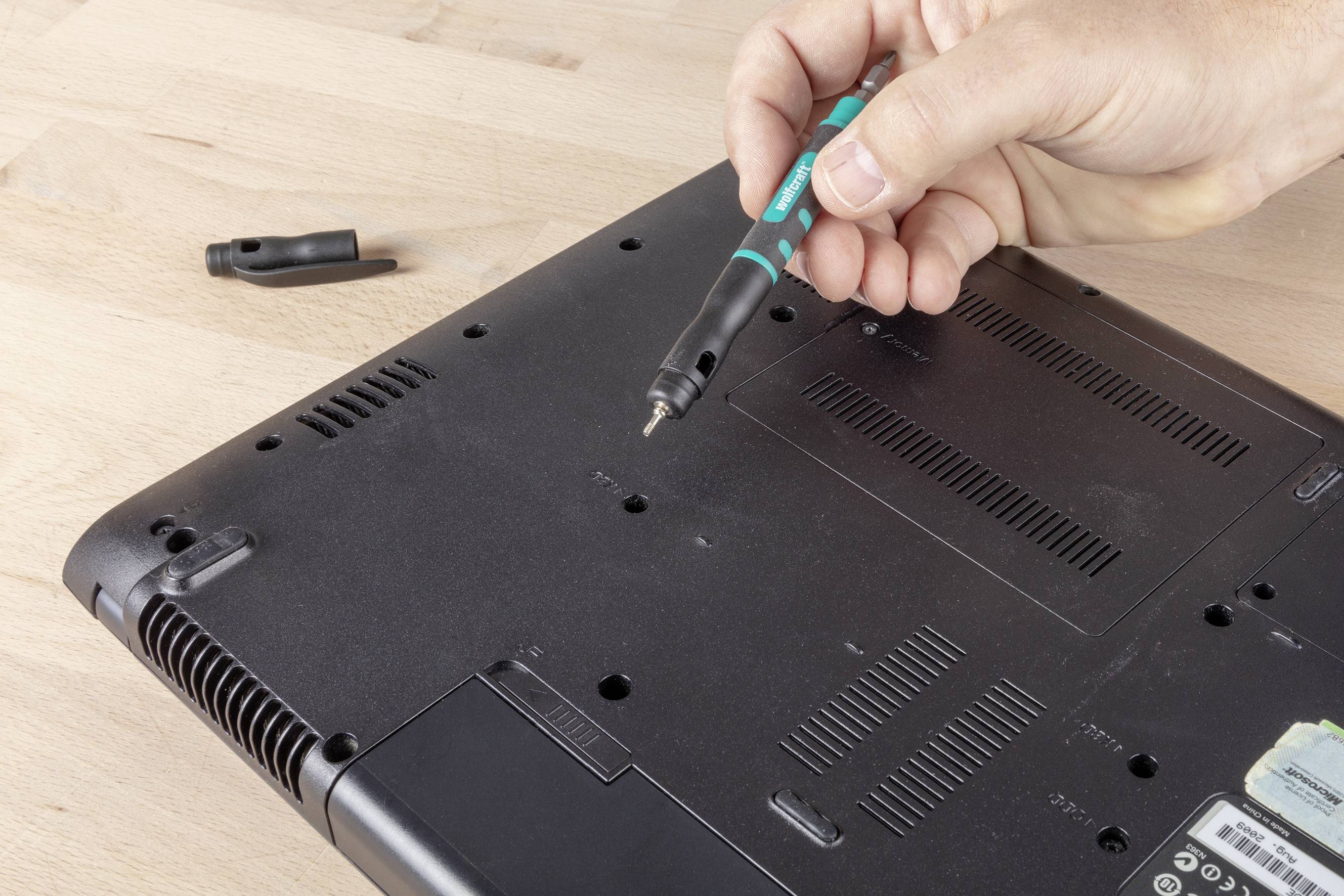 Eine Hand hält einen Schraubenzieher und entfernt eine Schraube aus der Rückseite eines elektronischen Geräts auf einem Holztisch.