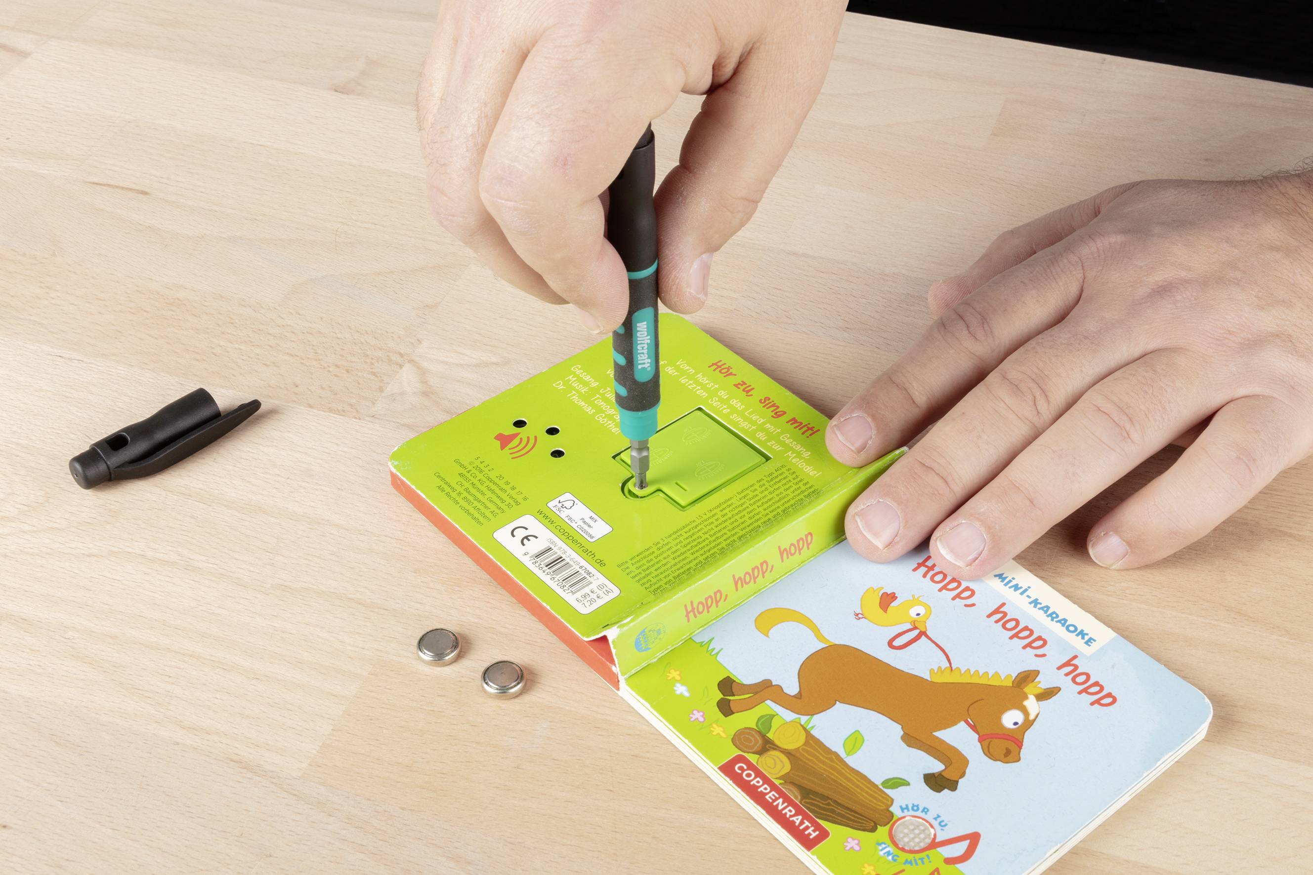 Eine Person arbeitet mit einem kleinen Buch und einem Schraubendreher auf einem Tisch, daneben liegen lose Batterien und ein Stiftdeckel.