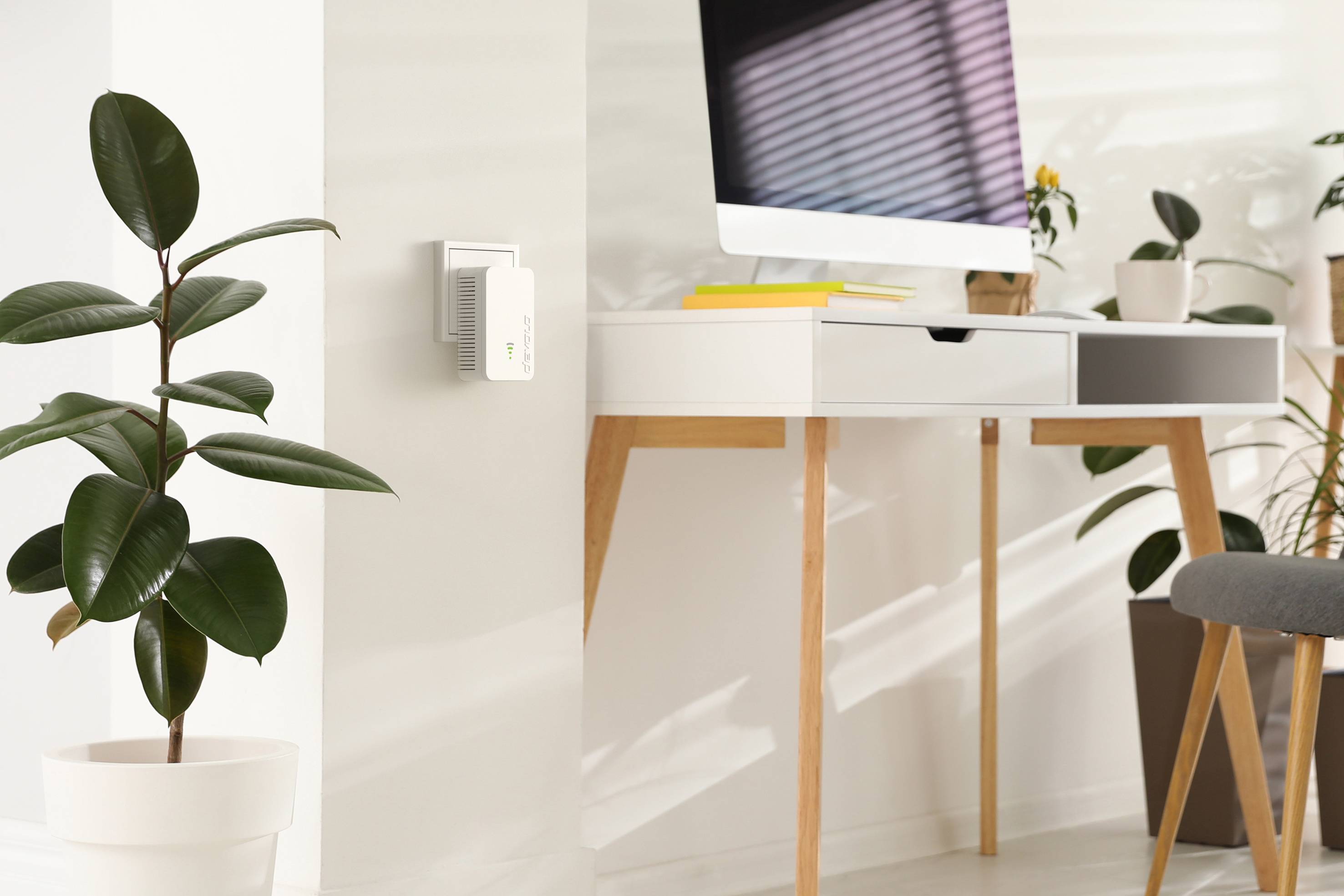 Moderner Arbeitsbereich mit einem hellen Holztisch, Computer und Zimmerpflanzen. Ein Gerät an der Wand zeigt ein grünes Licht.